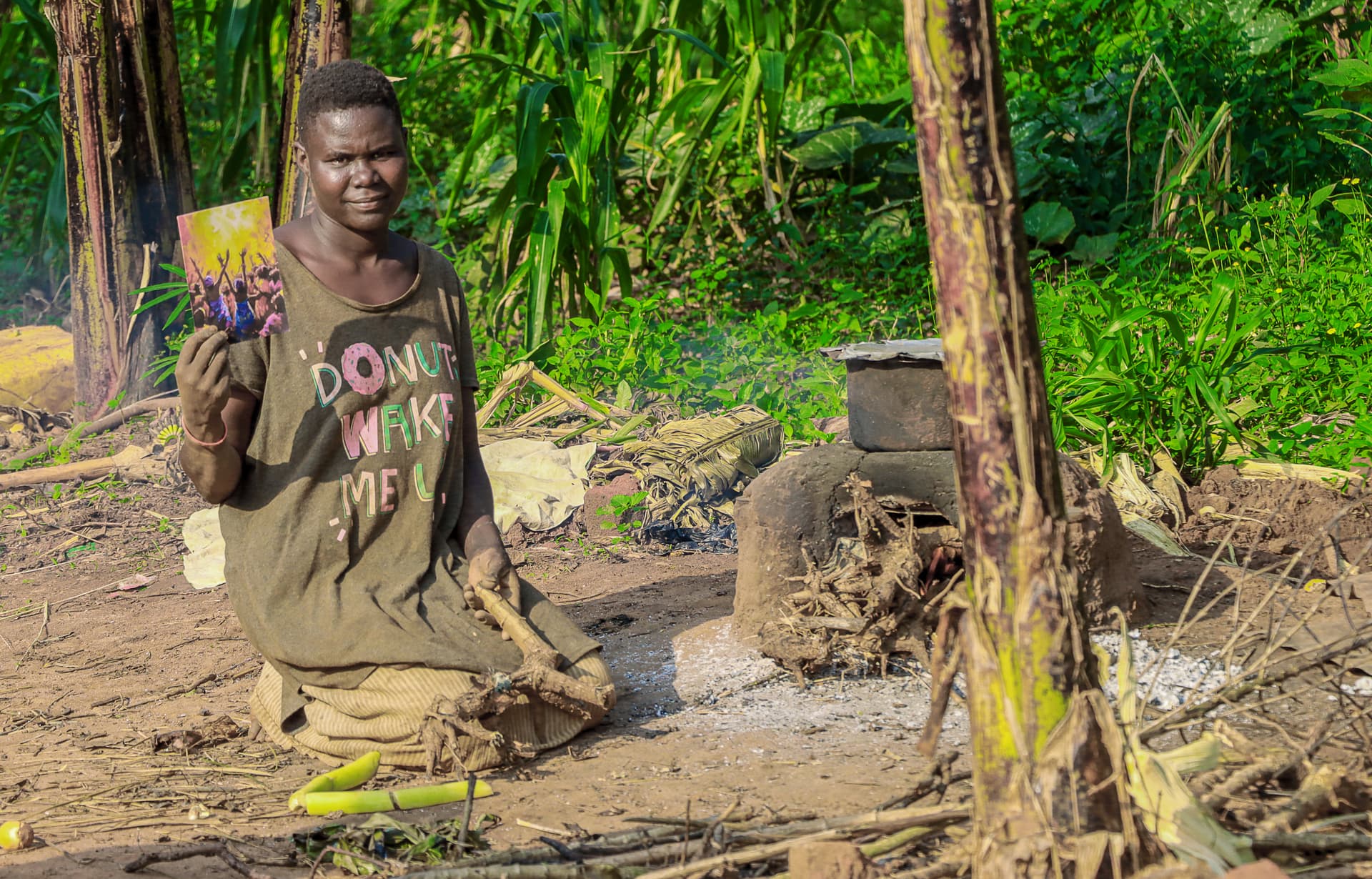 A woman, likely Mama Marvin, crouches tending an outdoor cooking fire beside a mud house.