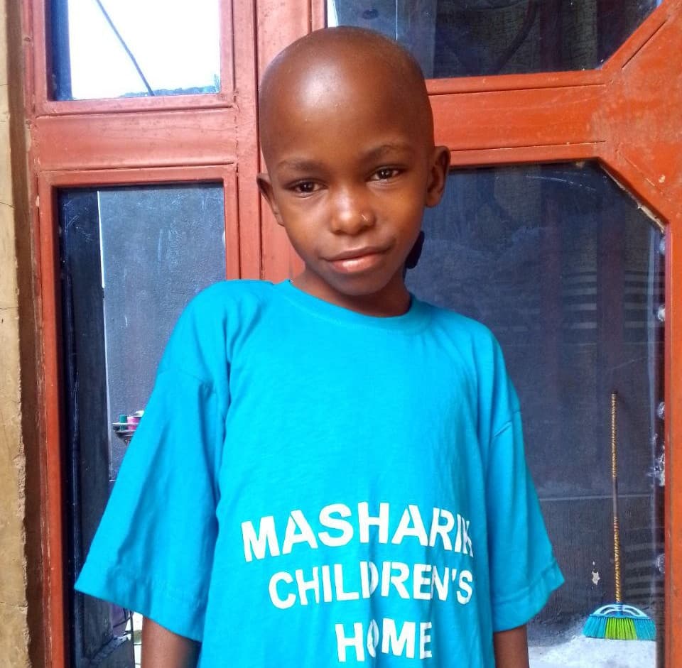A boy wearing a teal 'Mashariki Children's Home' t-shirt stands beside a red door.