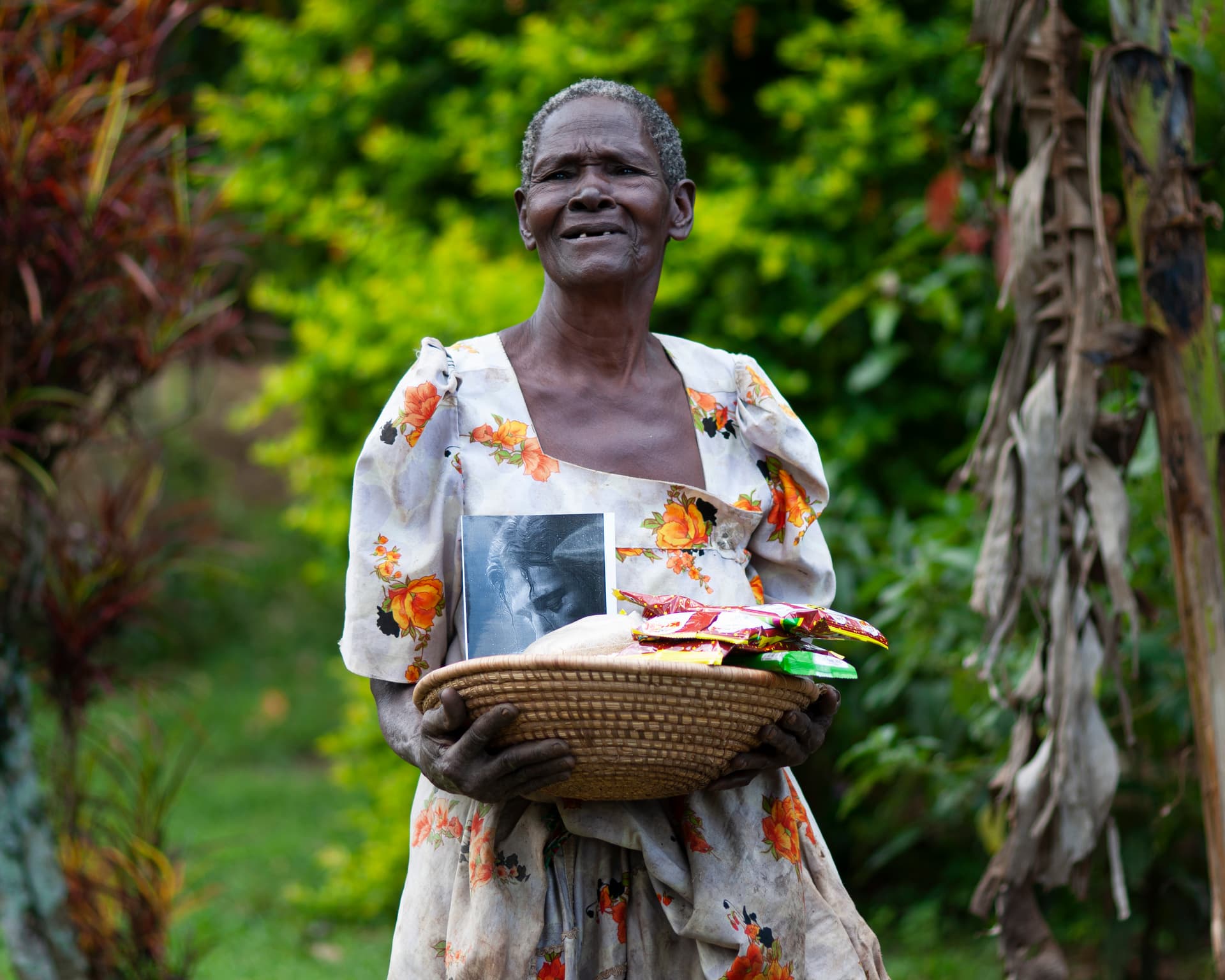 An elderly woman stands outdoors holding a woven basket filled with food gift packages, smiling among green trees.