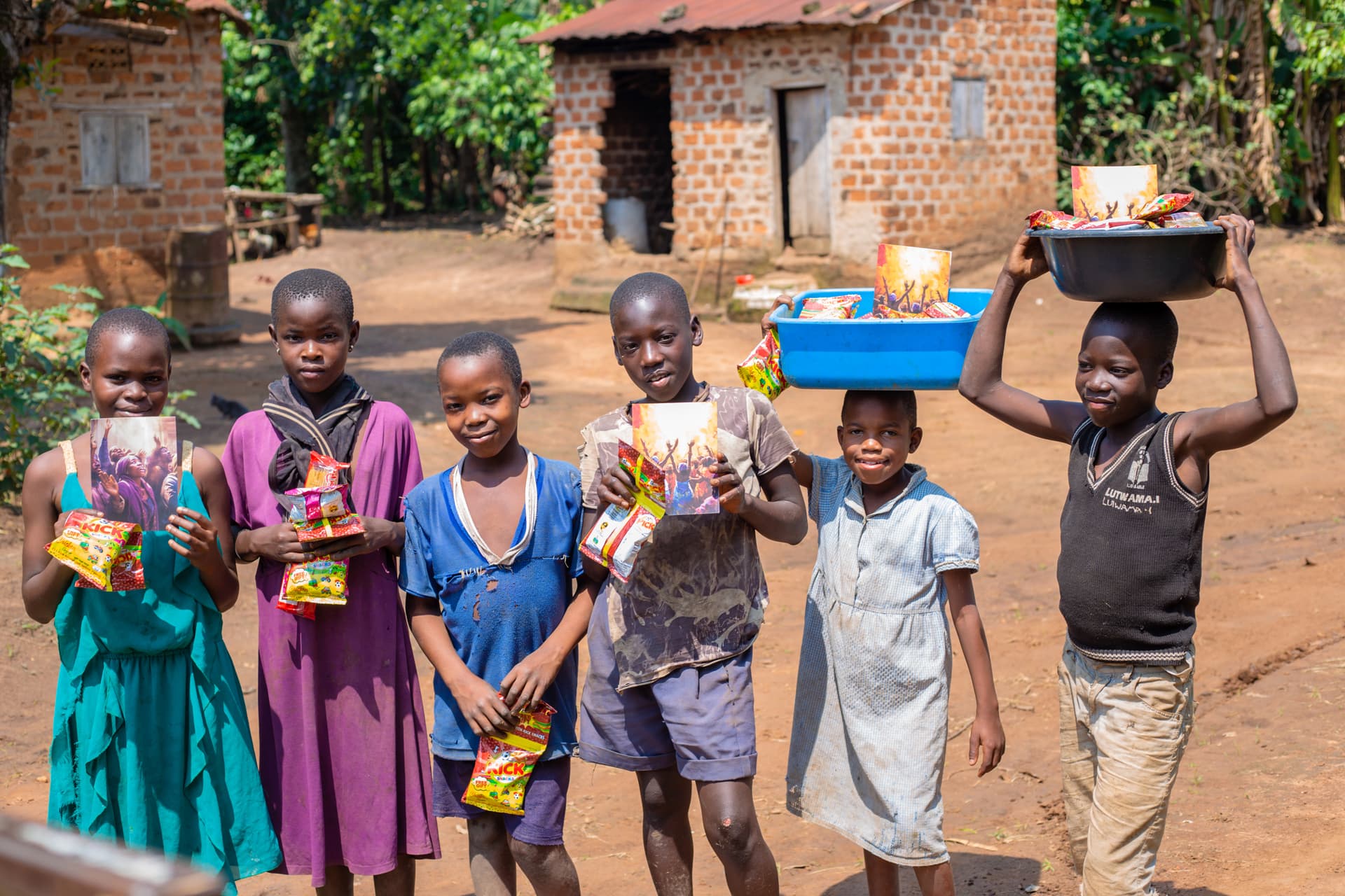 A group of children stand along a dirt path holding food gift items near brick houses, one boy balancing a tray on his head.