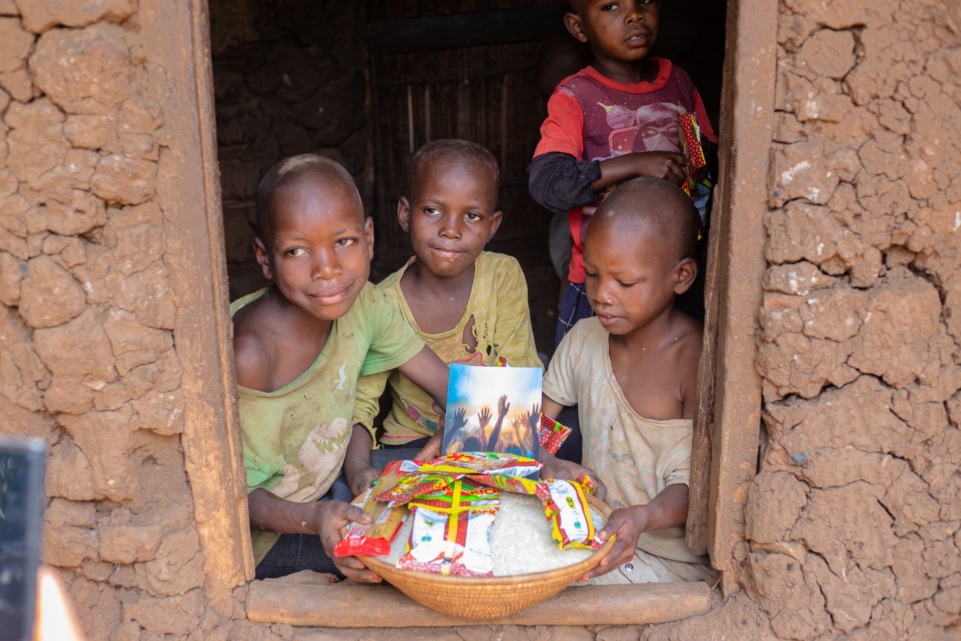 Several children stand in the doorway of a mud-brick house holding a basin of food packages and a printed photo card.
