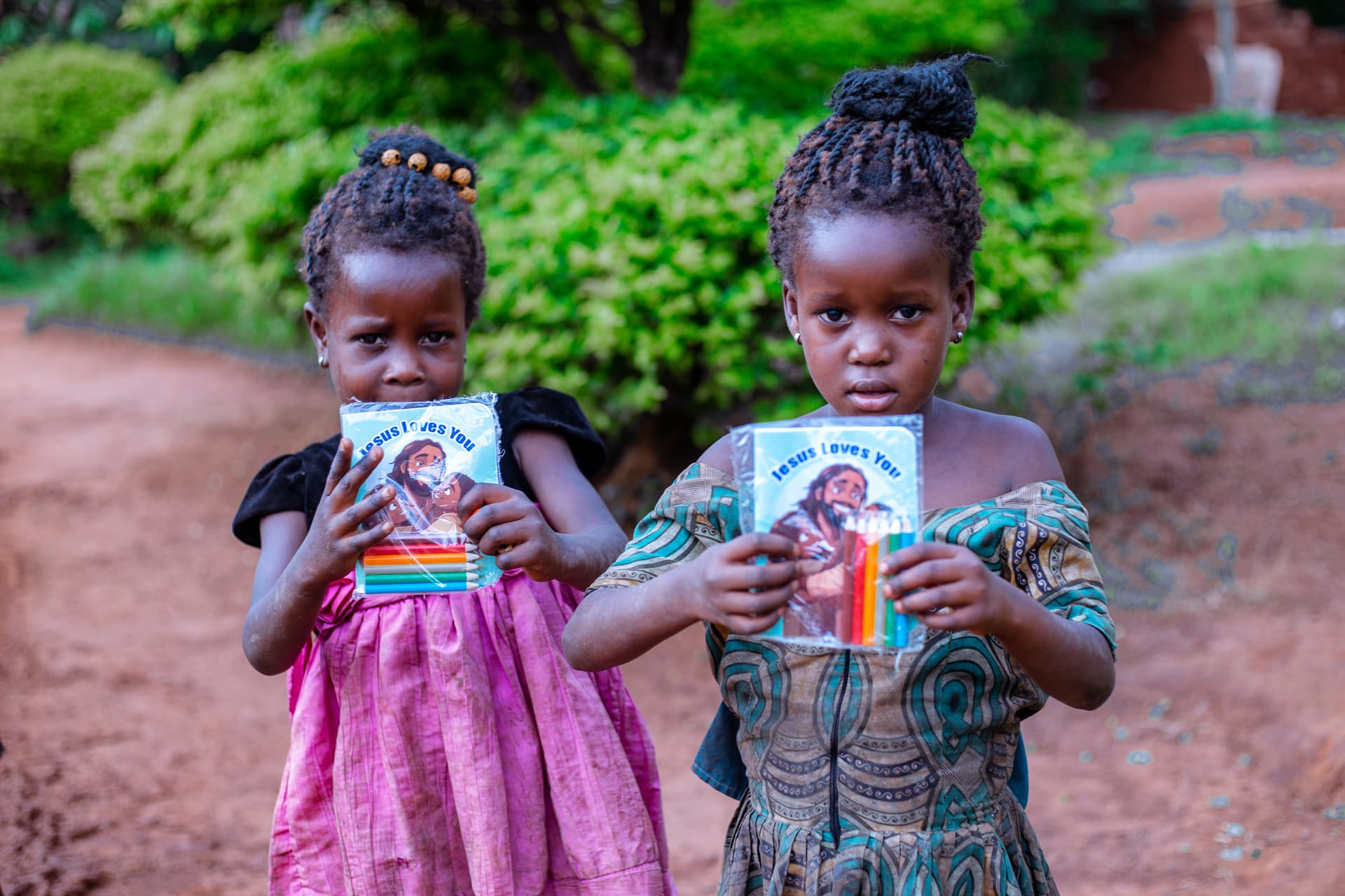 Two young girls stand on a dirt path holding children's booklets, one wearing a pink dress.