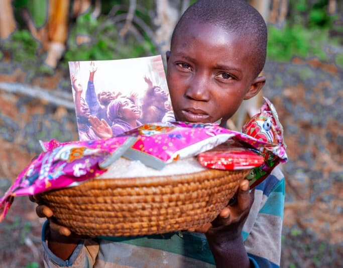 A boy holds a woven basket filled with colorful food gift packages and a booklet, standing outdoors.