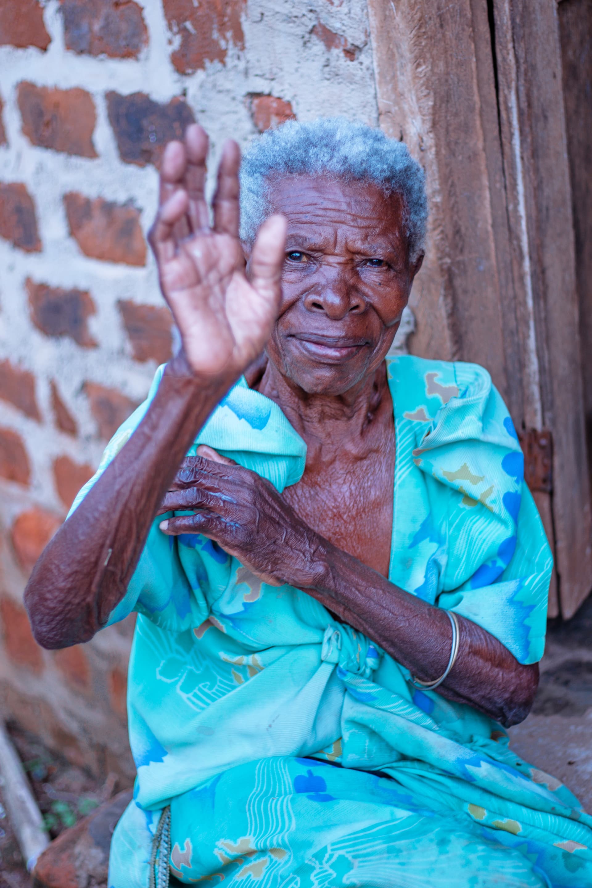 An elderly woman in a teal dress sits outside a brick house, raising her hand in greeting.