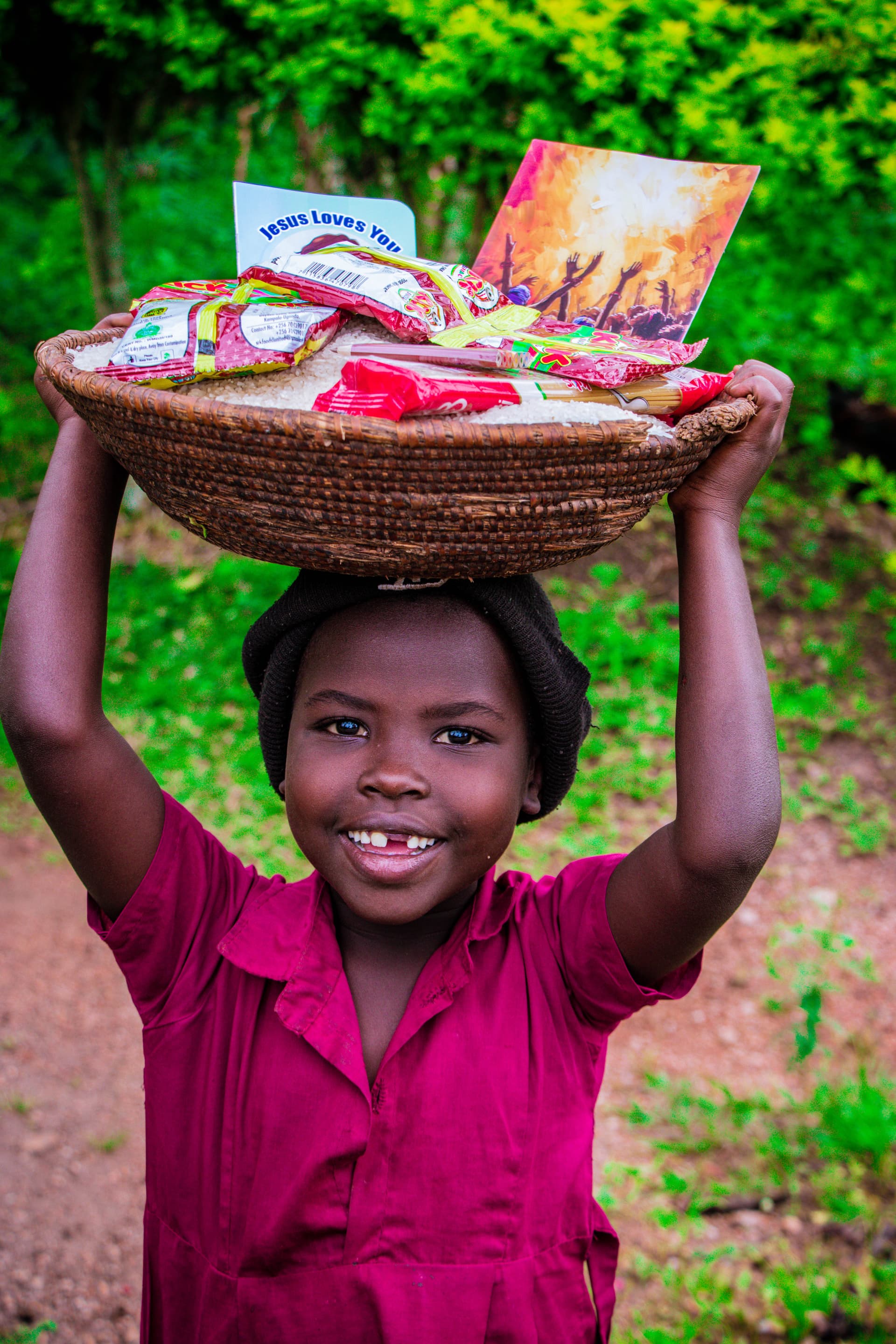 A young girl smiles while carrying a basket of food gift packets on her head, likely Lillian.