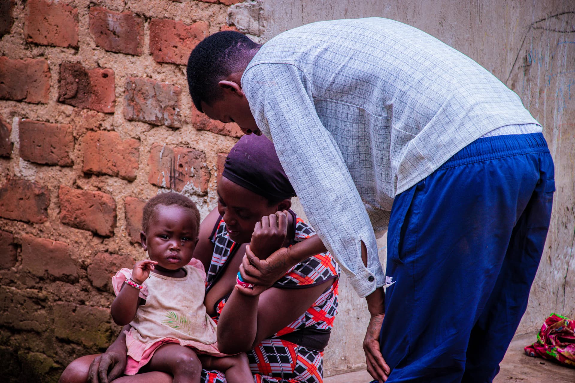 A man in a white shirt bends down to pray with two children outside a brick wall.