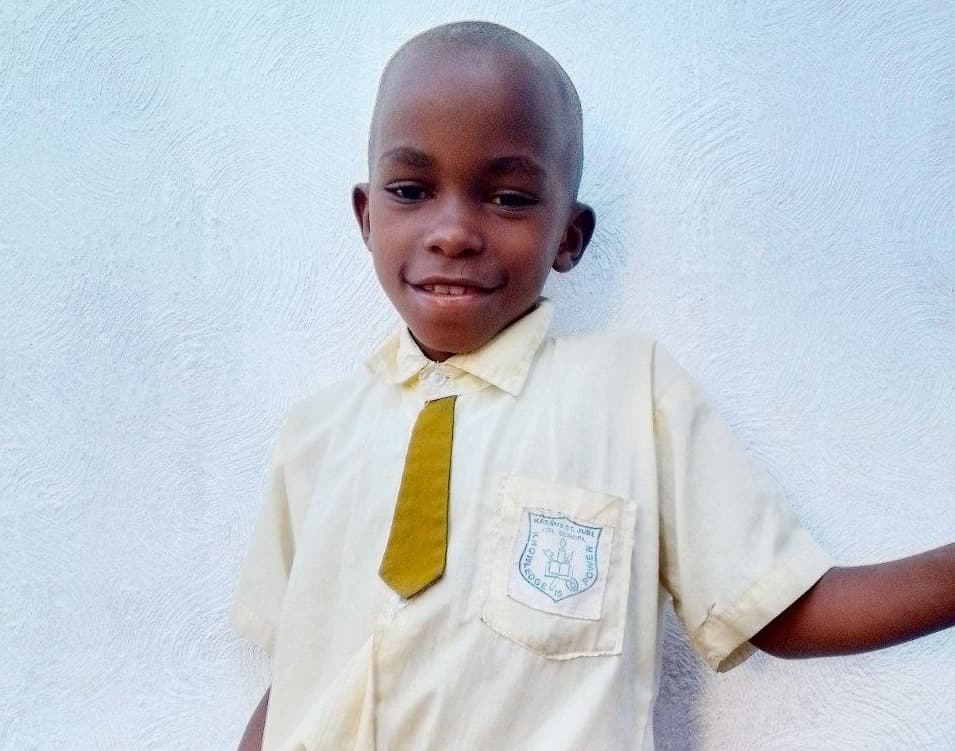 Boy in a cream school uniform shirt and dark tie, smiling against a white wall.