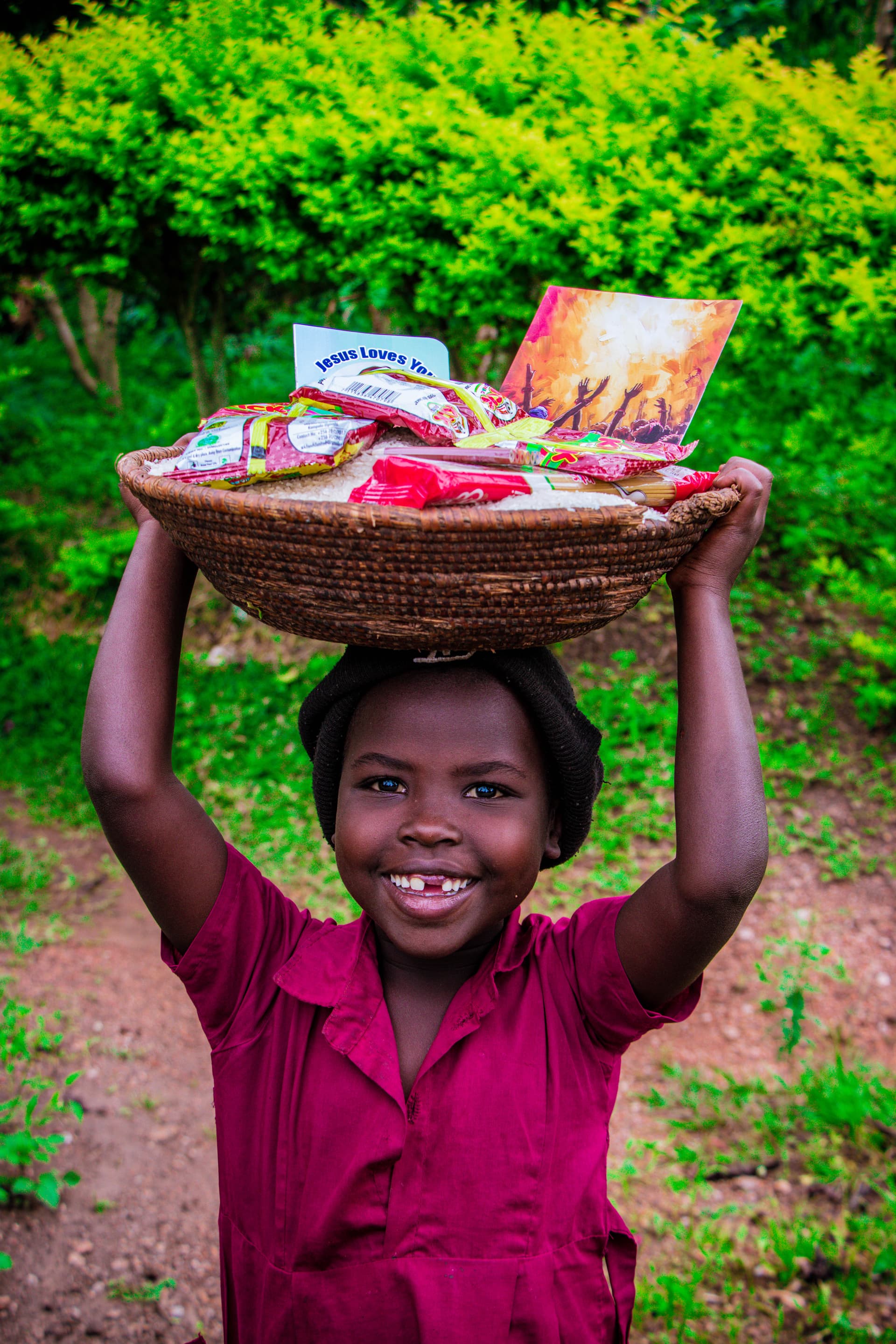 Girl in a red dress balances a basket of food gift packets and a gospel tract on her head, smiling.