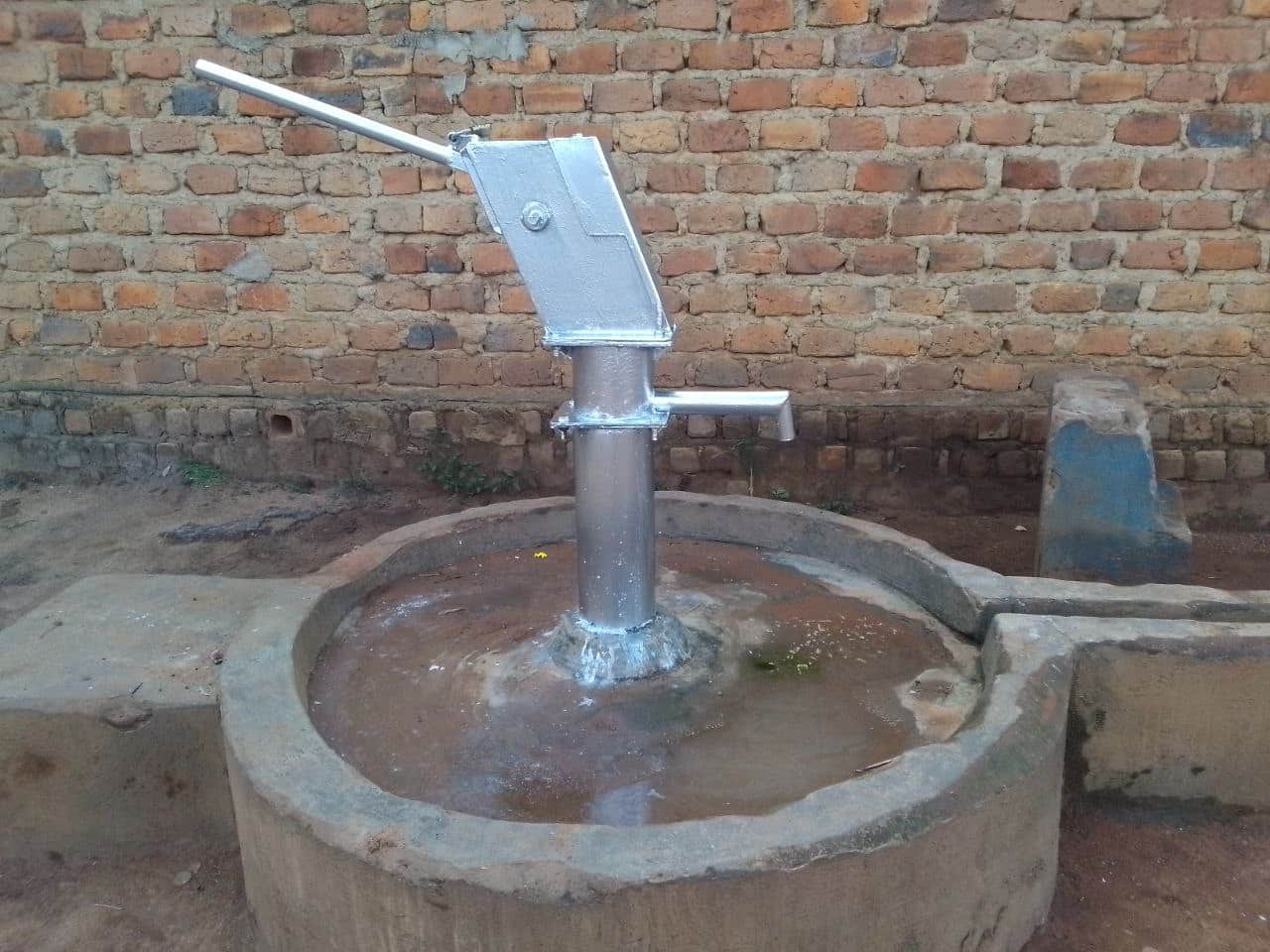 Hand-pump borehole well with a concrete apron, known locally as the Jesus Well, beside a brick wall.
