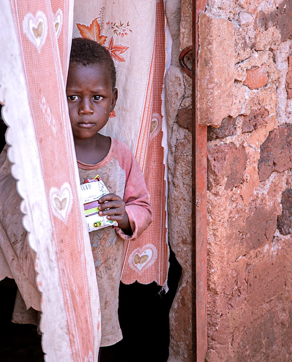 Child peeking out from behind a pink curtain in a doorway, holding a small item.