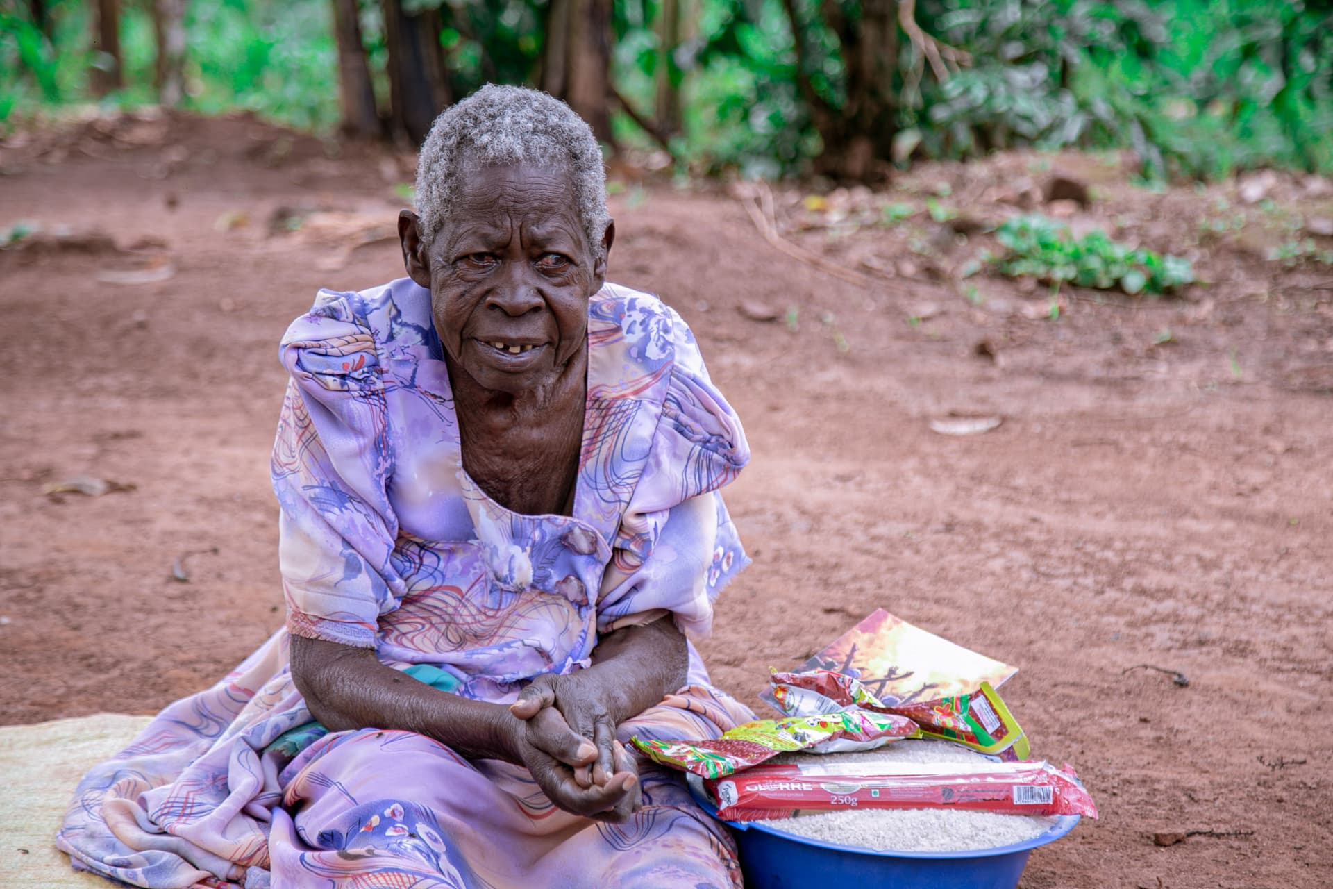 Elderly woman in a purple floral dress sitting outdoors with a basin of food items.