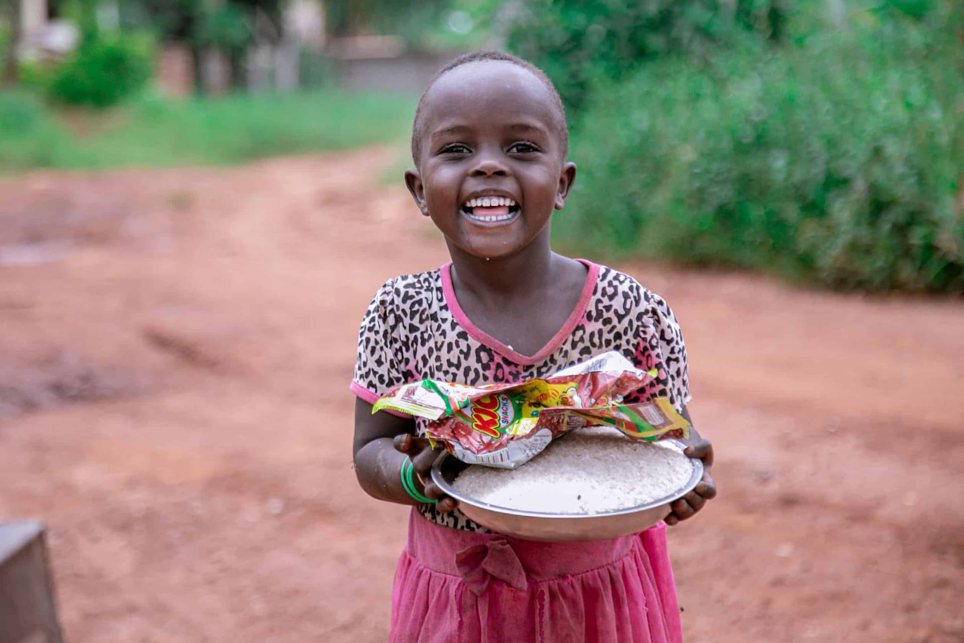 Smiling girl in a pink skirt holding a plate of food items on a dirt road.