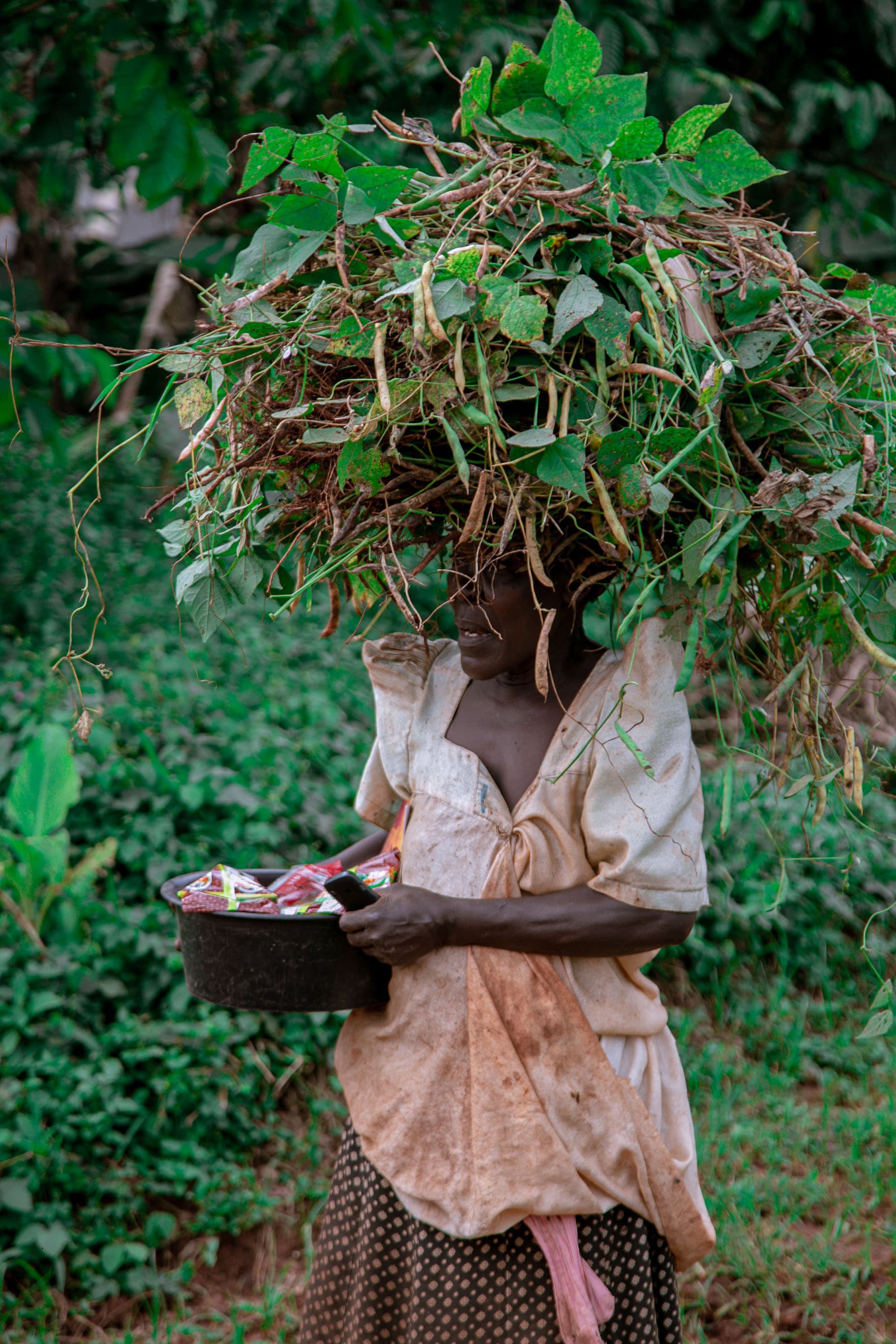 Woman balancing a bundle of leafy greens on her head while carrying a food basin.