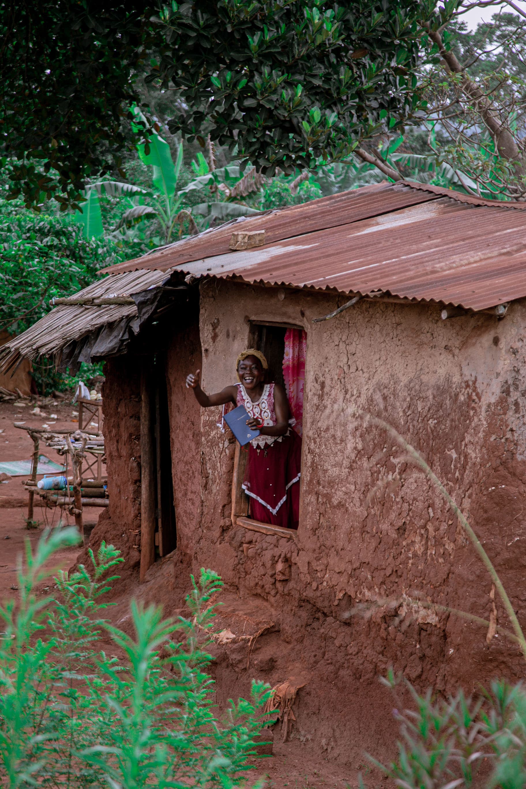 Woman in a headscarf and red skirt standing in the doorway of a mud-brick house with a tin roof.