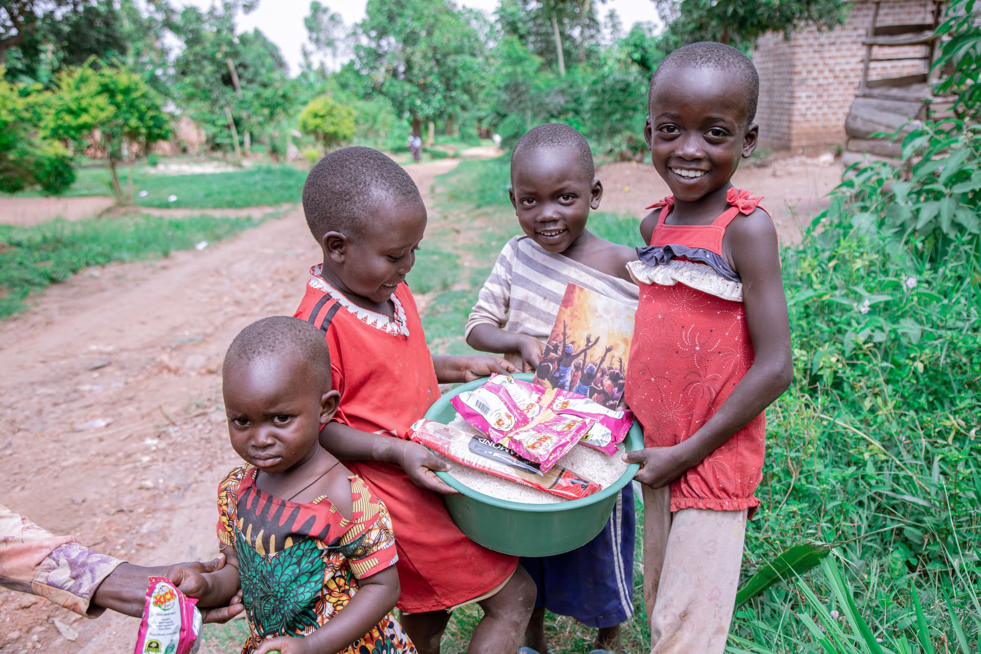 A group of four children stand together outdoors holding food gift packets, one wearing a red dress.