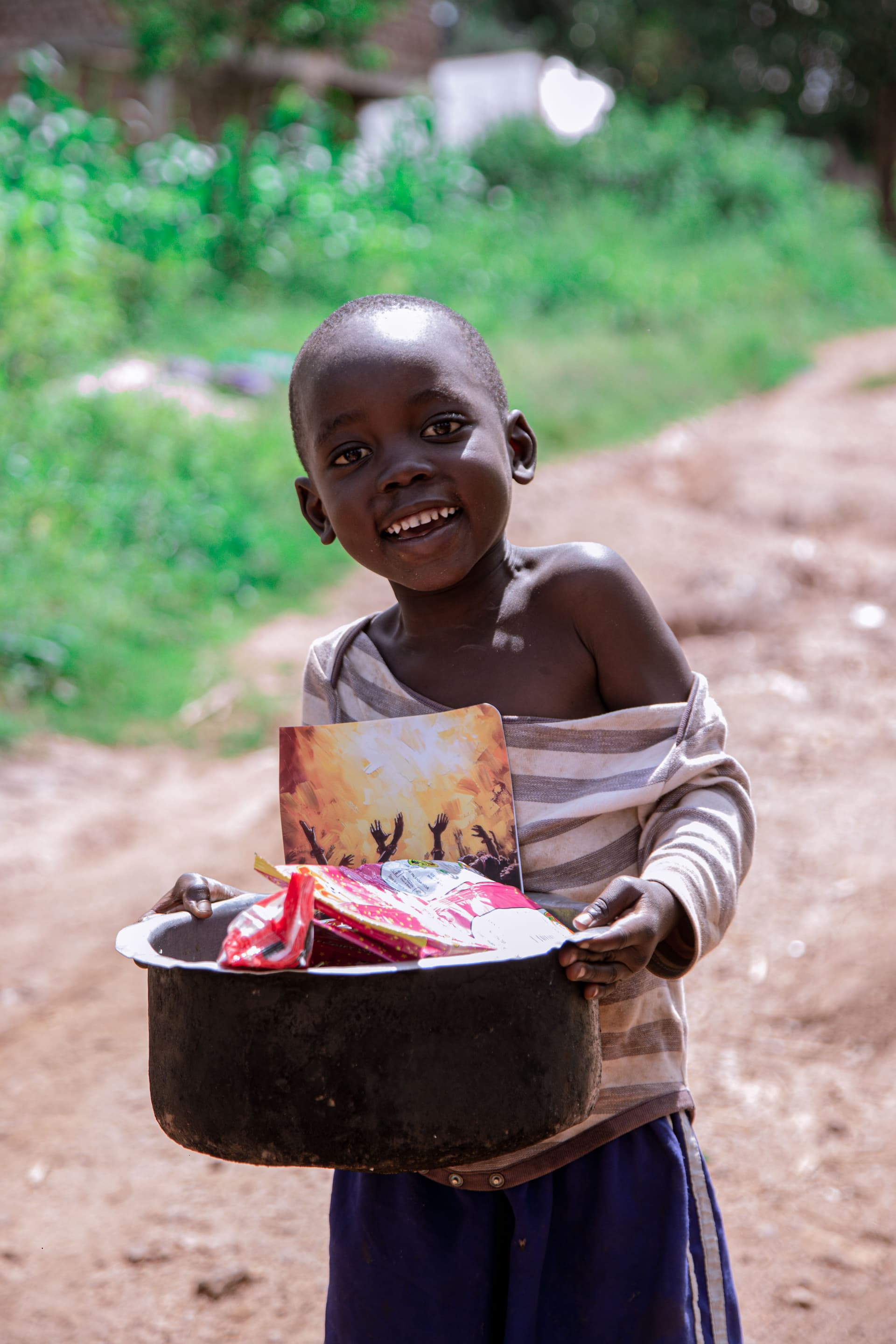 A shirtless boy smiles while holding a pot filled with food gift packets on a dirt road.