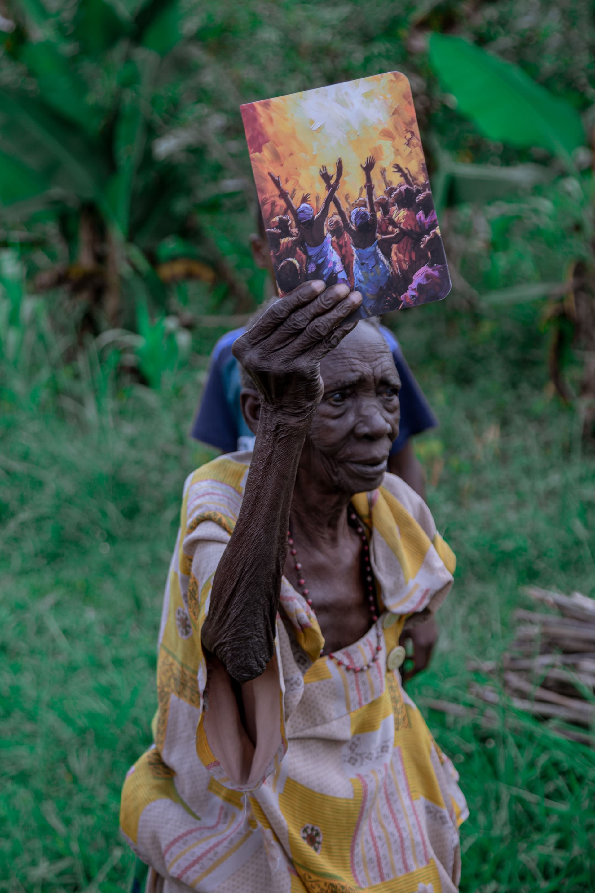 Elderly Ugandan woman balancing a food gift package on her head while standing in tall grass.