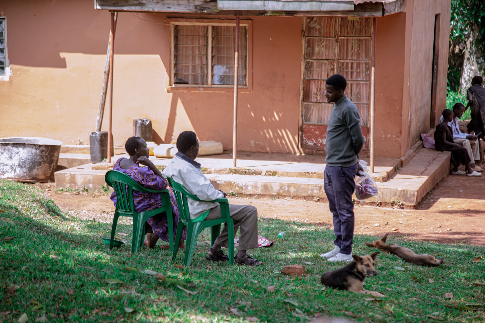 Elderly men sit in plastic chairs outside a peach-colored house while another man stands nearby, a dog resting on the grass.