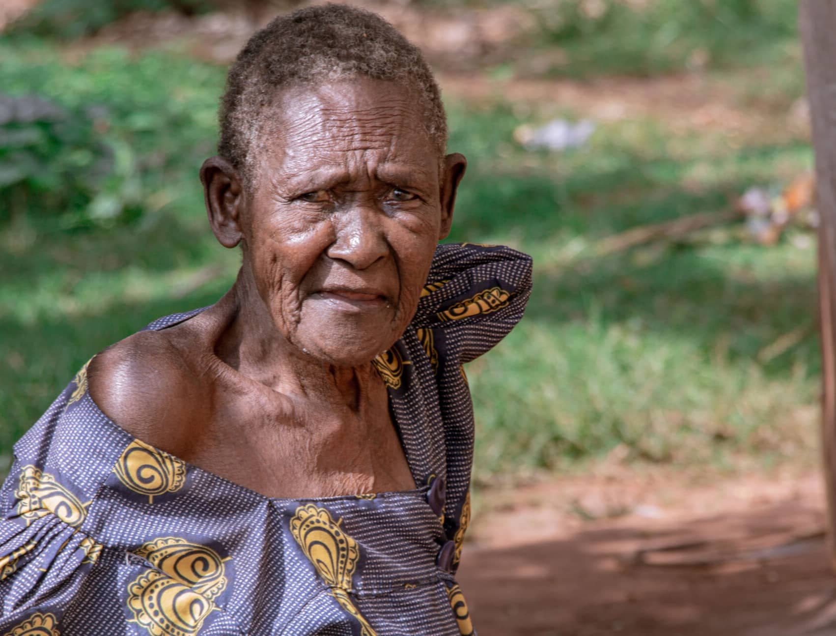 An elderly woman stands outdoors in a green, leafy setting.