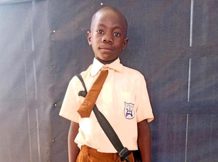 Yahaya stands against a gray wall wearing a white school uniform shirt and tie.