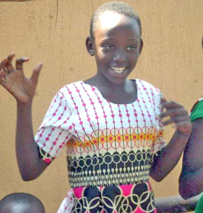 Gloria, a young girl in a colorful patterned dress, laughs with her hands raised outdoors.