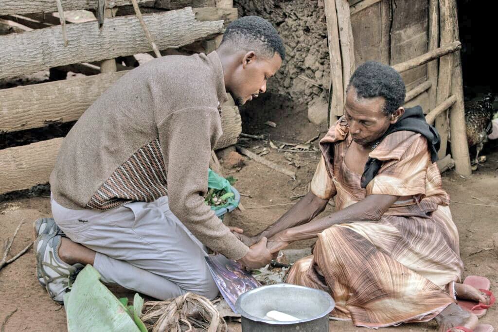 A young man helps wash the hands of an elderly seated woman outside a thatched hut.