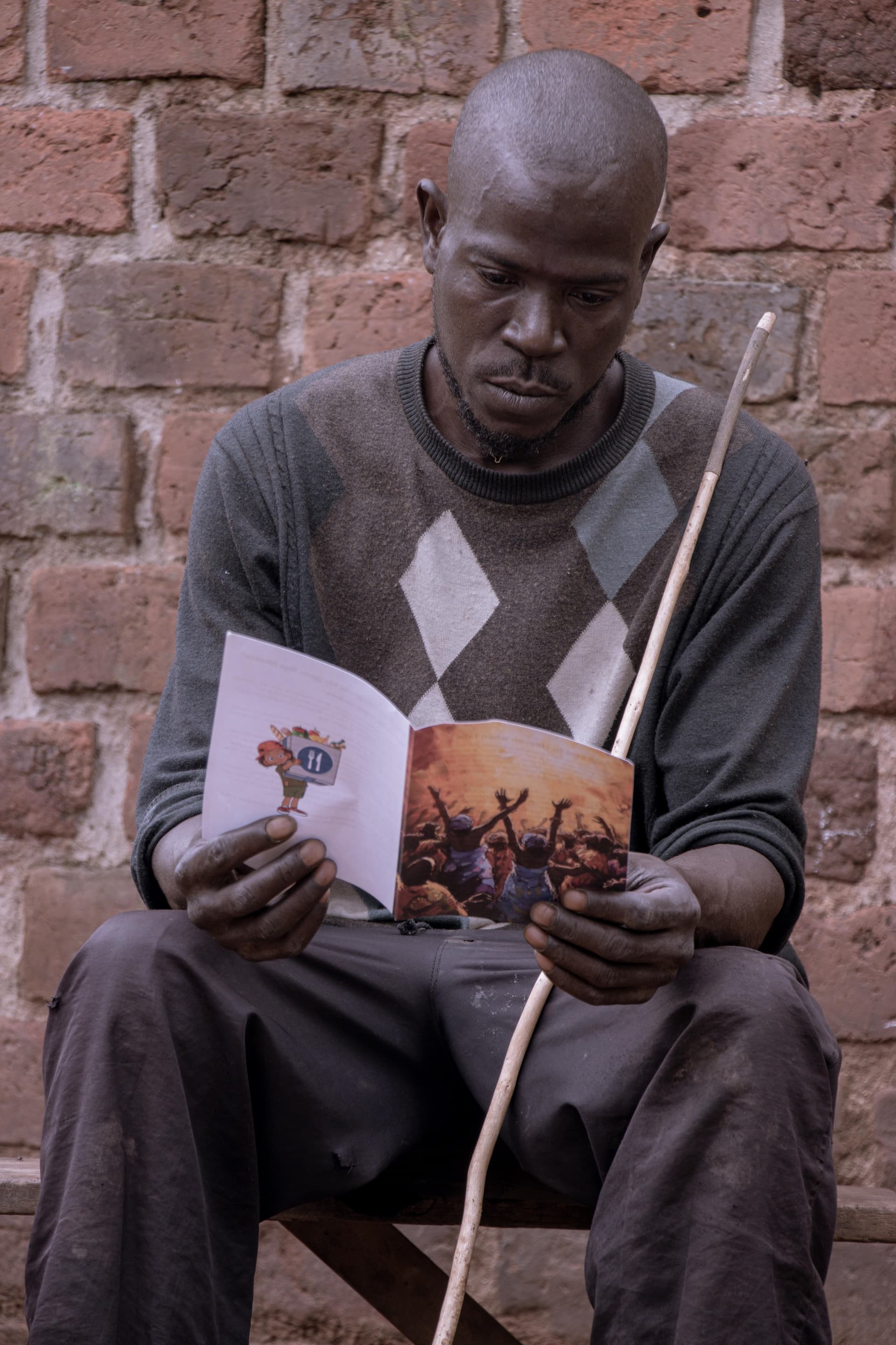 A man, likely Sserunjogi Paul, reads a gospel tract while seated against a brick wall with a walking stick beside him.