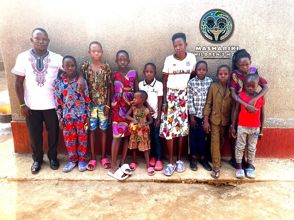 A group of children and a man pose together outdoors in front of a school building, some wearing traditional African print clothing.