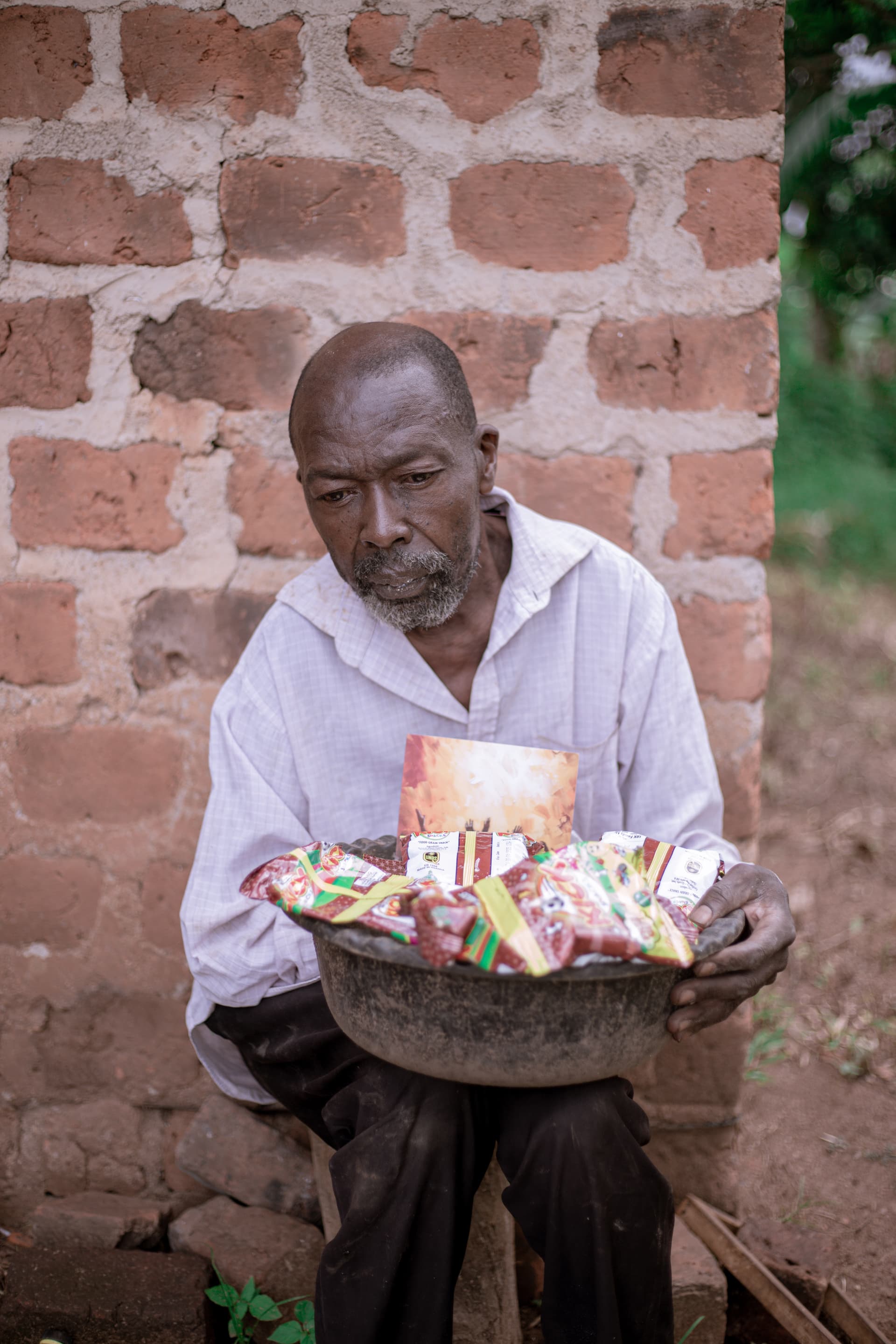 An elderly man in a white shirt holds a basin of snack packets on his lap outdoors.