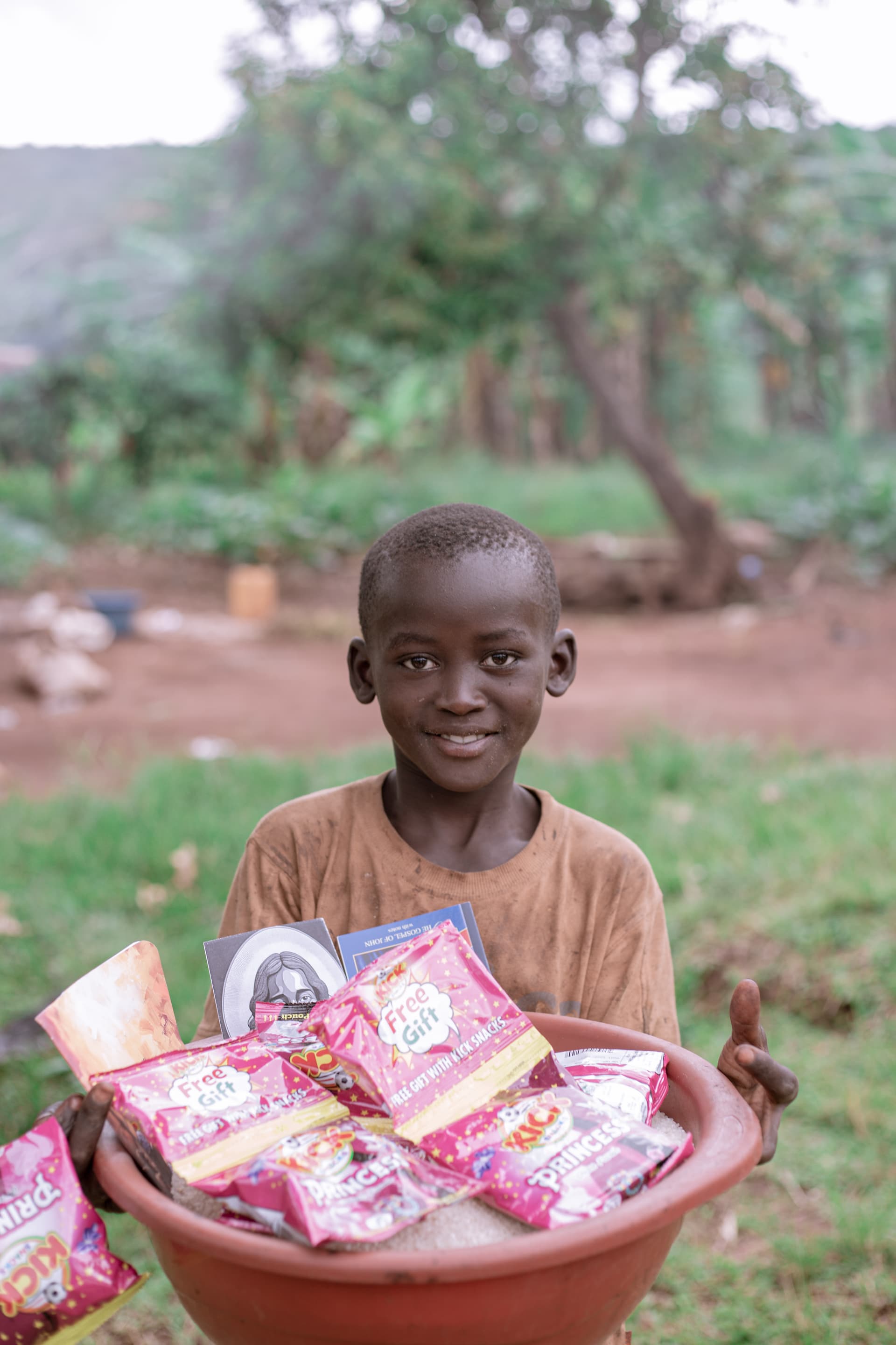 A boy smiles while holding a red basin filled with snack packets, standing on a grassy area.