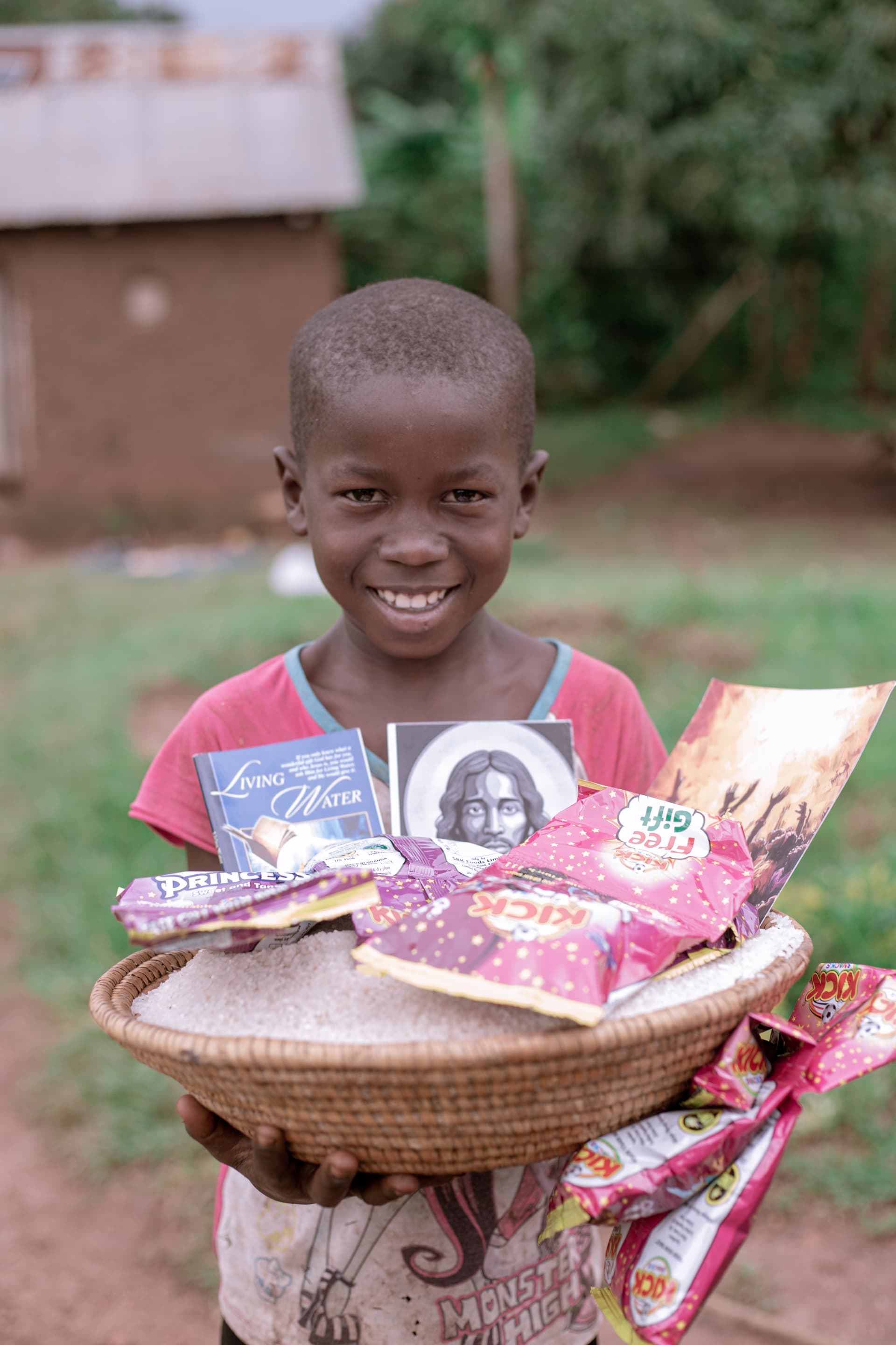 A boy smiles while holding a basket of snack packets and a photograph, standing outdoors.