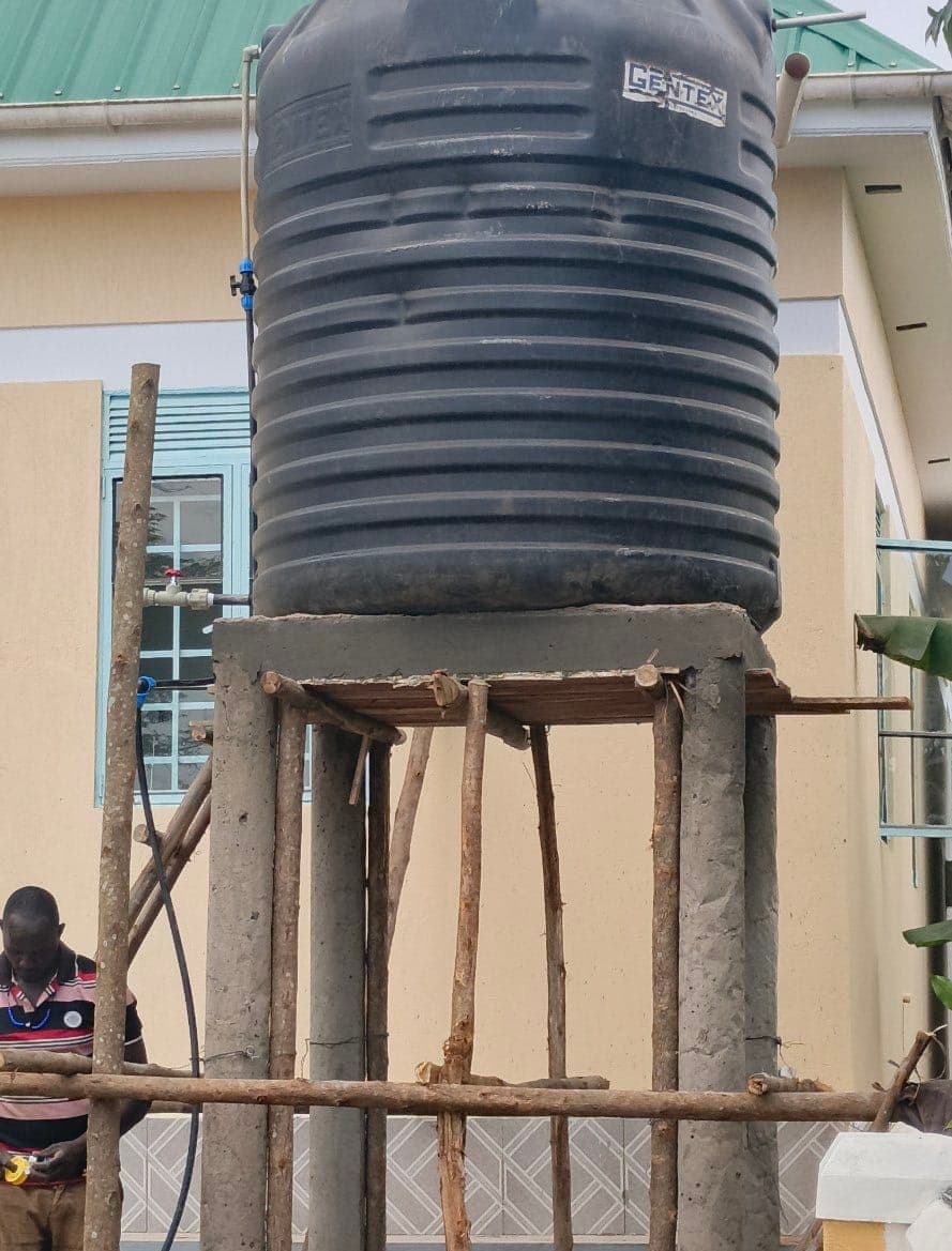 A large water storage tank on a wooden and concrete stand outside the Miracle Medical Center building, a person seated nearby.