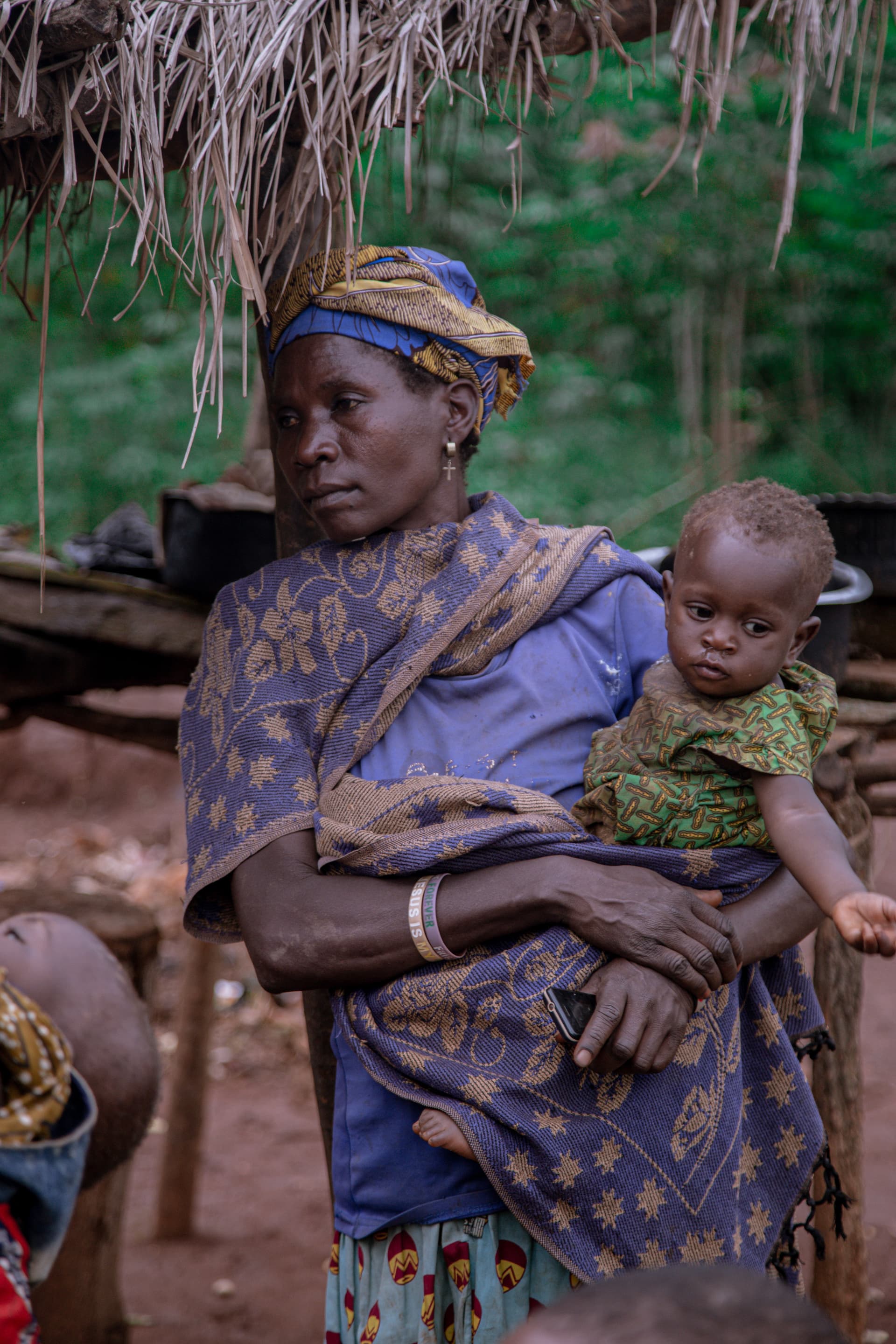 Woman in traditional dress holding a toddler on her lap, seated under a thatched-roof shelter.