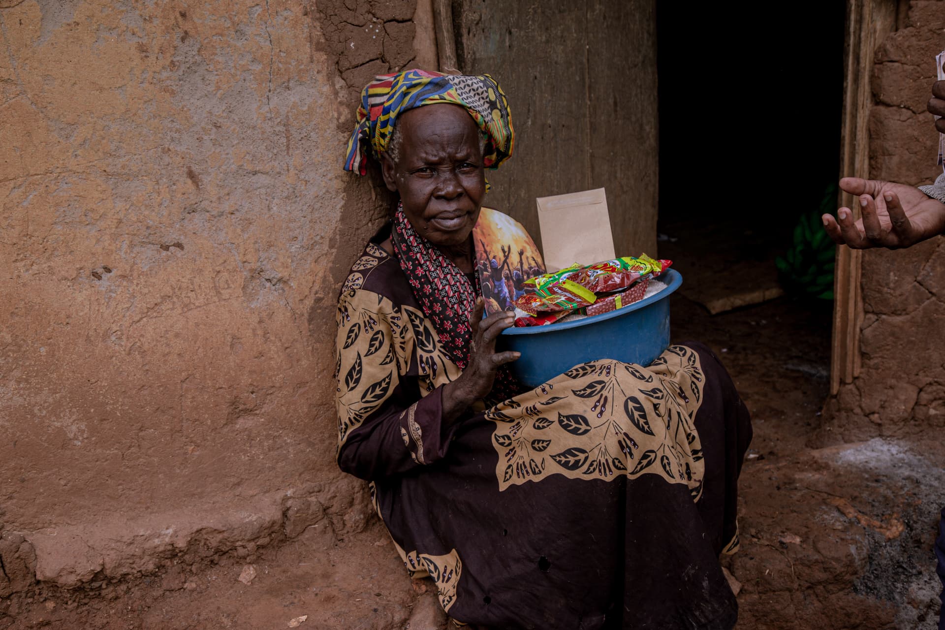 An elderly woman in a headscarf sits outside a mud-brick home holding a blue basin of Christmas food gifts.
