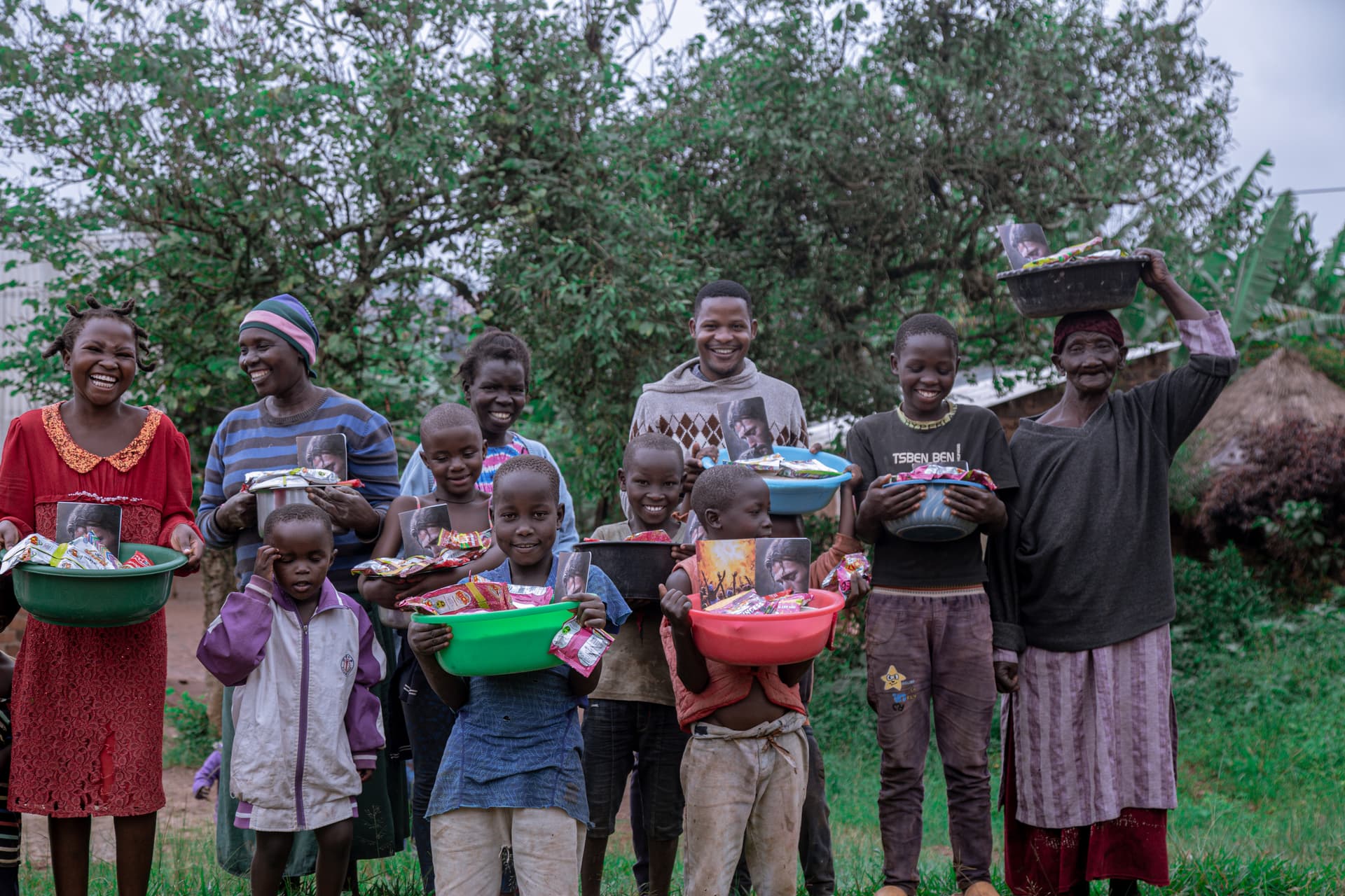 A group of children and a woman hold basins of food gifts, standing together outdoors among trees.
