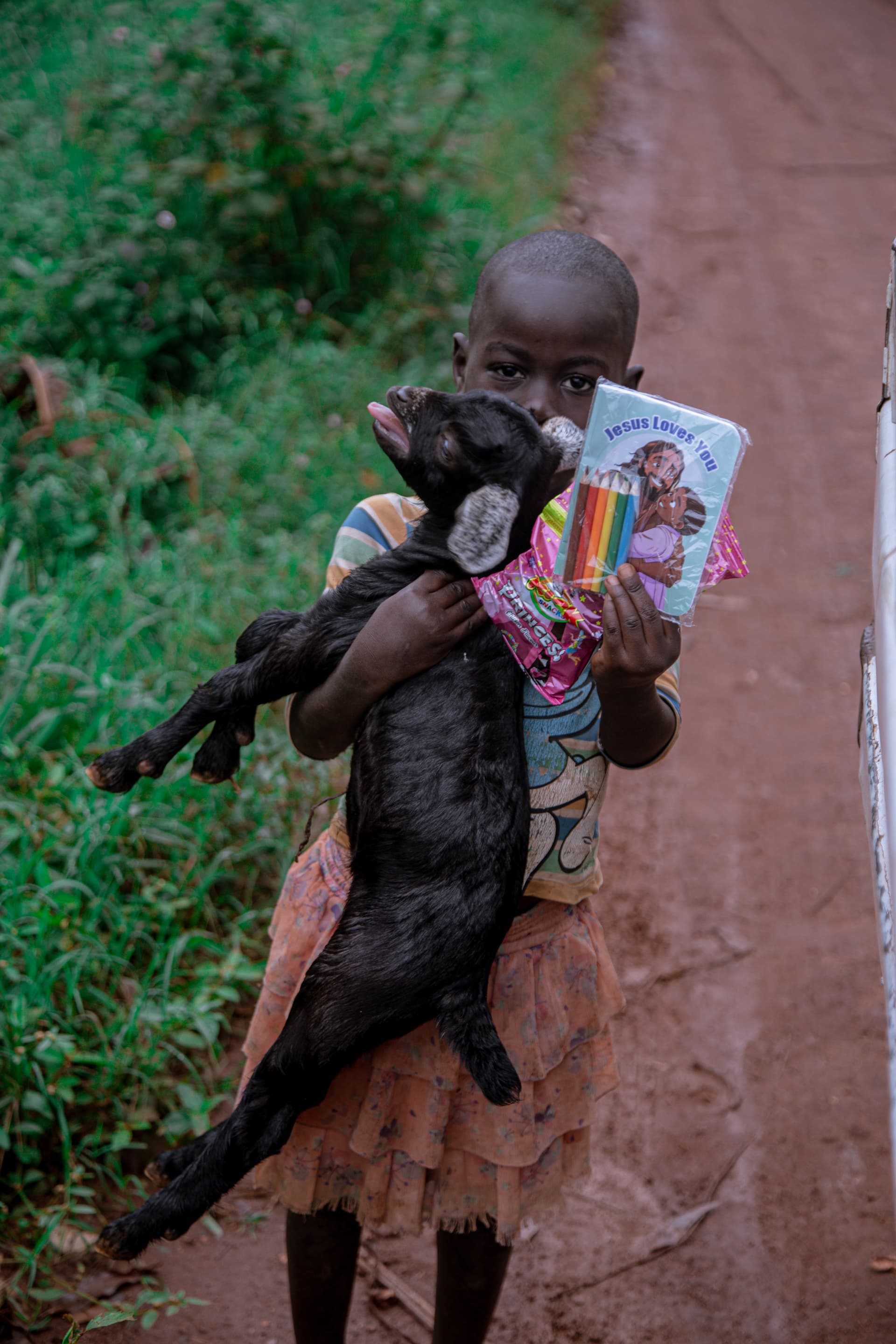 Leticia, a young girl, cradles her baby goat in her arms while holding a magazine, standing on a dirt path.