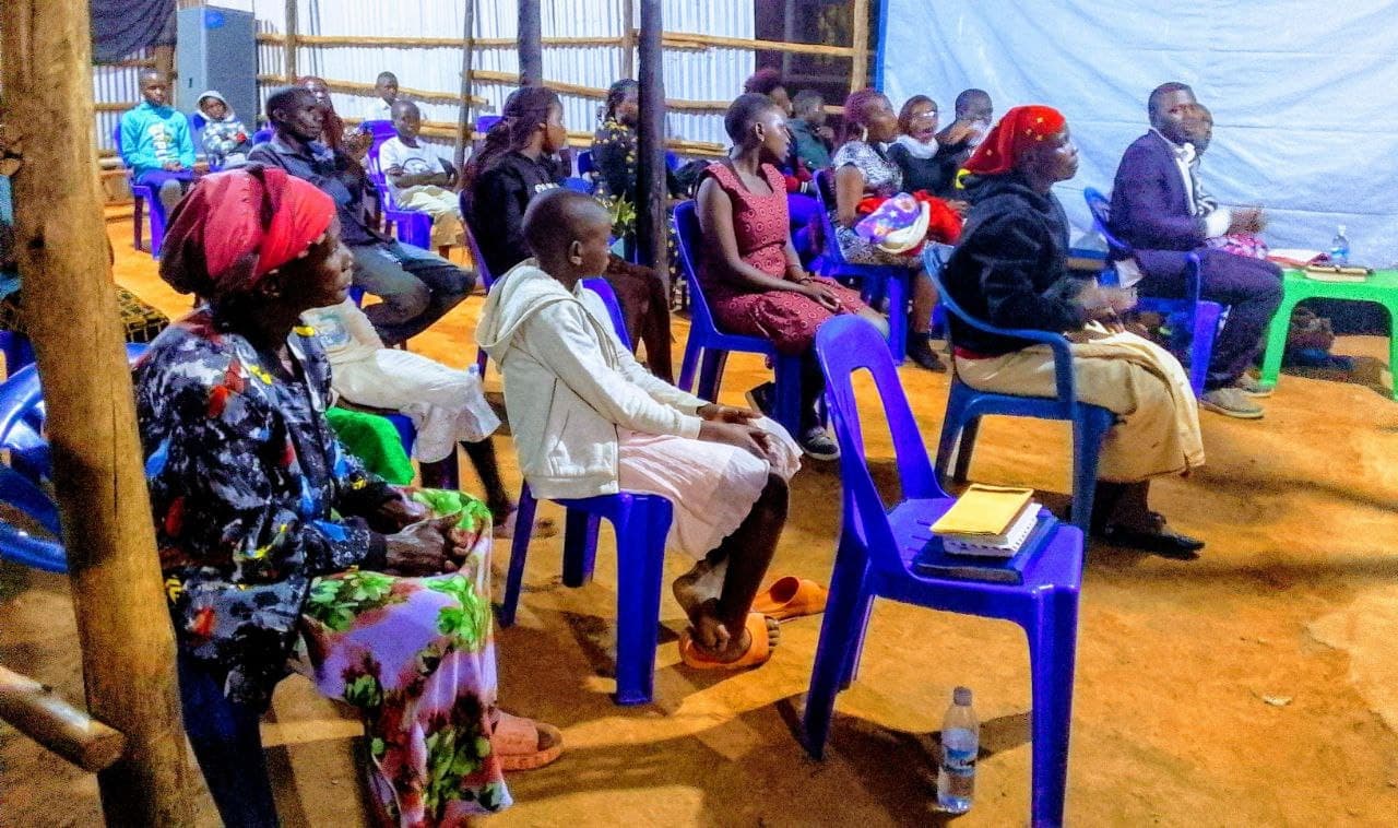 People seated in blue plastic chairs under a tent canopy at a year-end gathering, light filtering through the fabric.