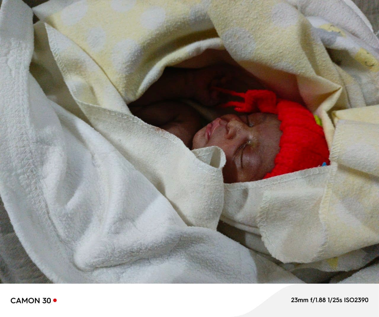Newborn baby wrapped in a white blanket with a red cap, sleeping.