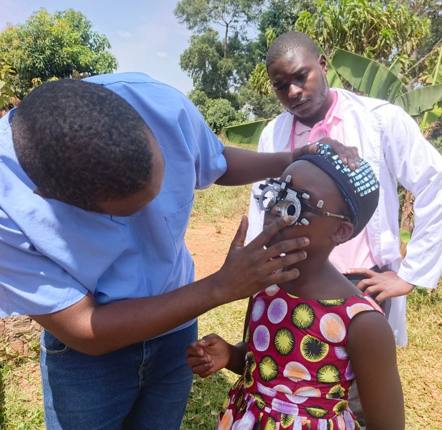 Man in a white coat helping a girl try on eyeglasses outdoors while another man assists.