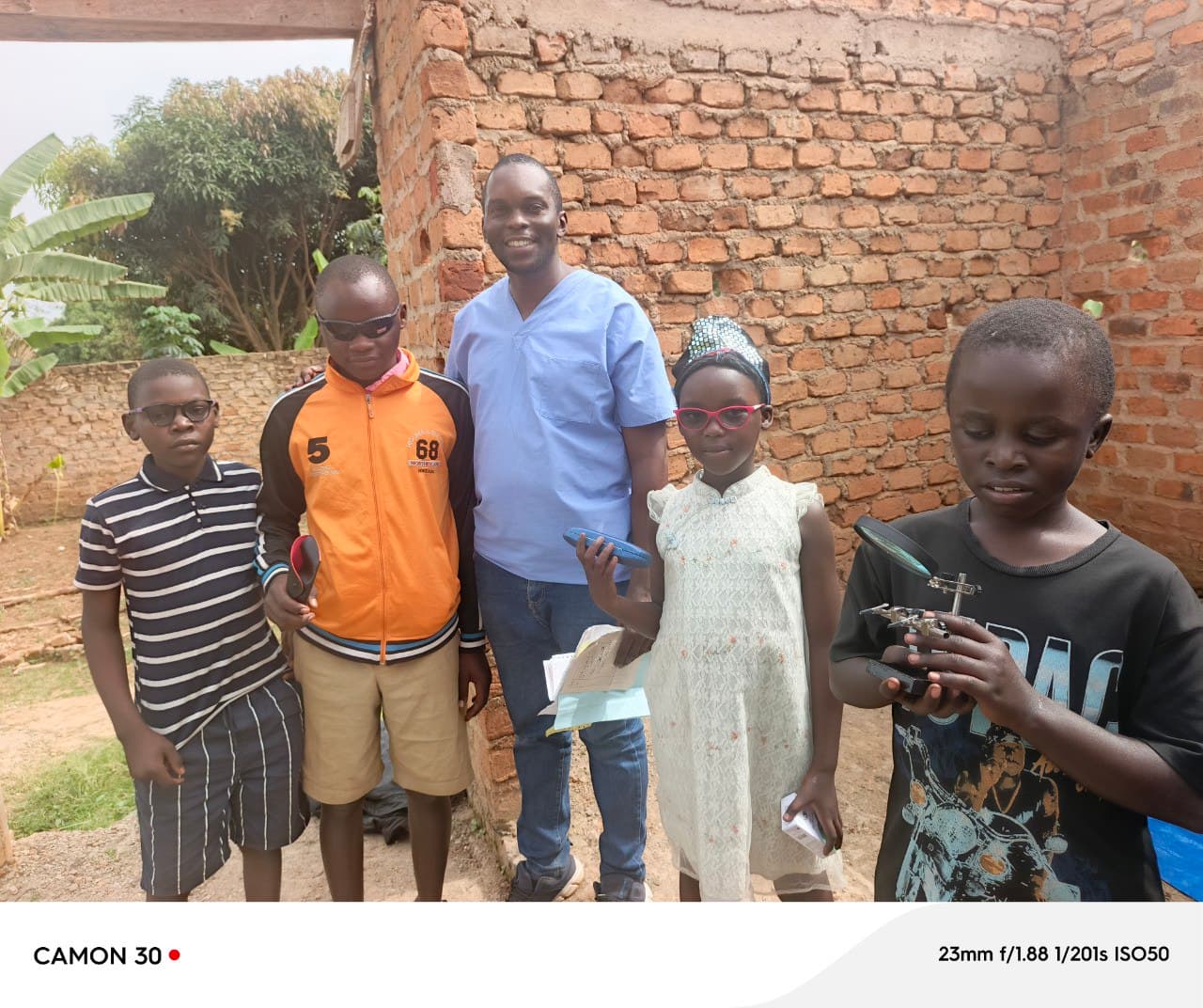 Group photo of four children and a man in a blue shirt standing outdoors against a brick wall.