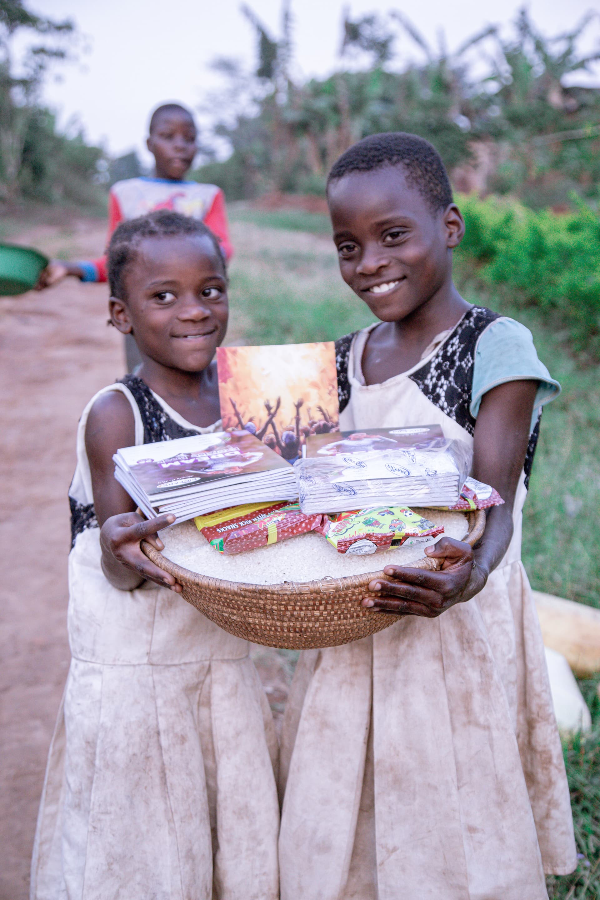 Two girls in traditional dresses smile while carrying a shared basket of goods in a rural setting.
