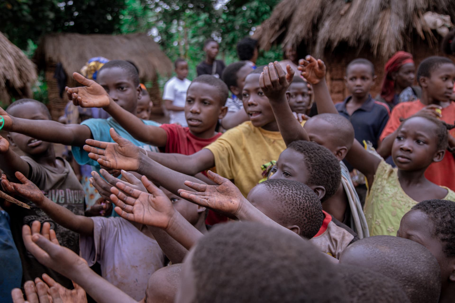 A crowd of children and adults reach their hands toward the camera at an outdoor gathering near thatched-roof huts.
