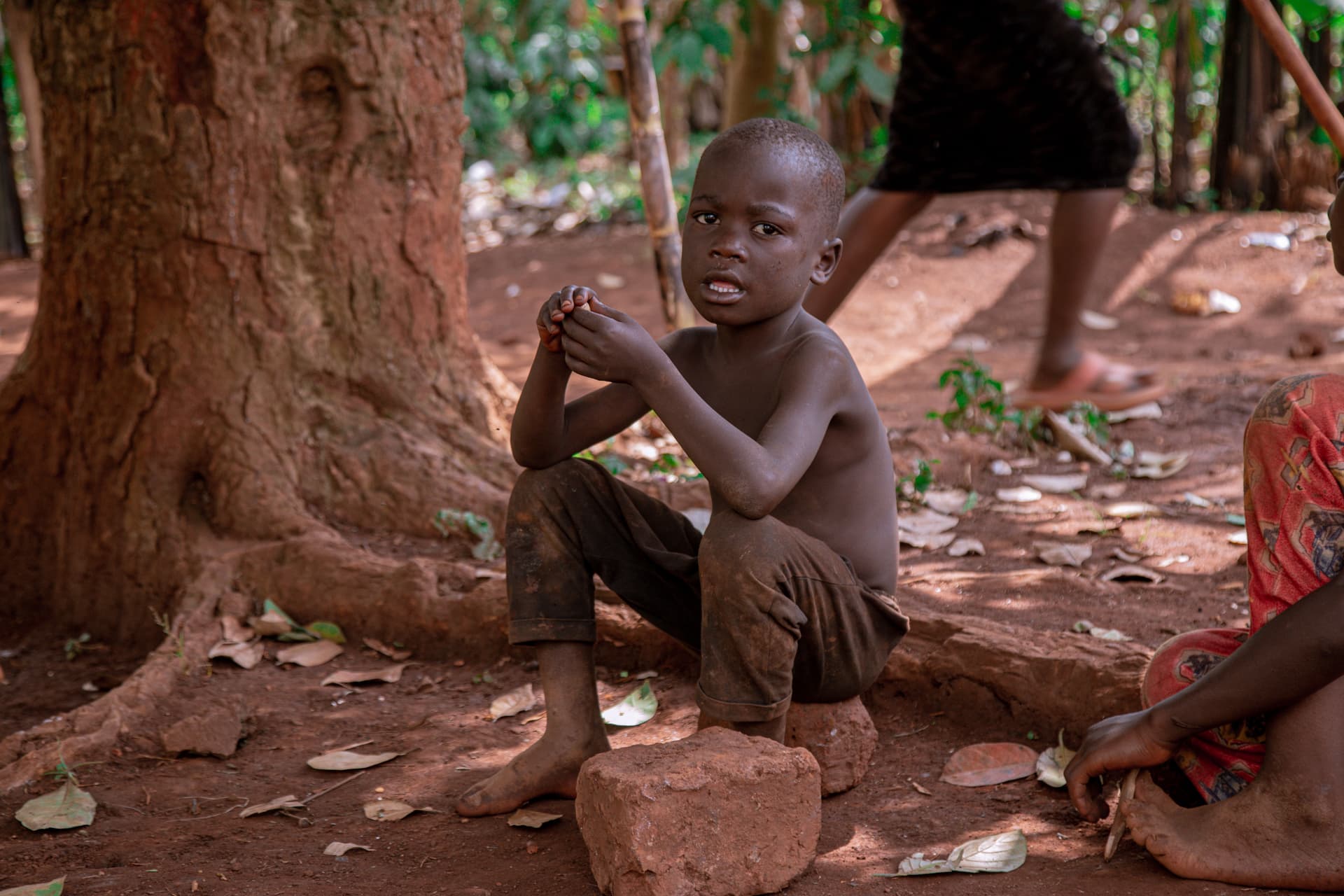 Boy sitting on the ground under a tree, bare feet, looking at the camera.