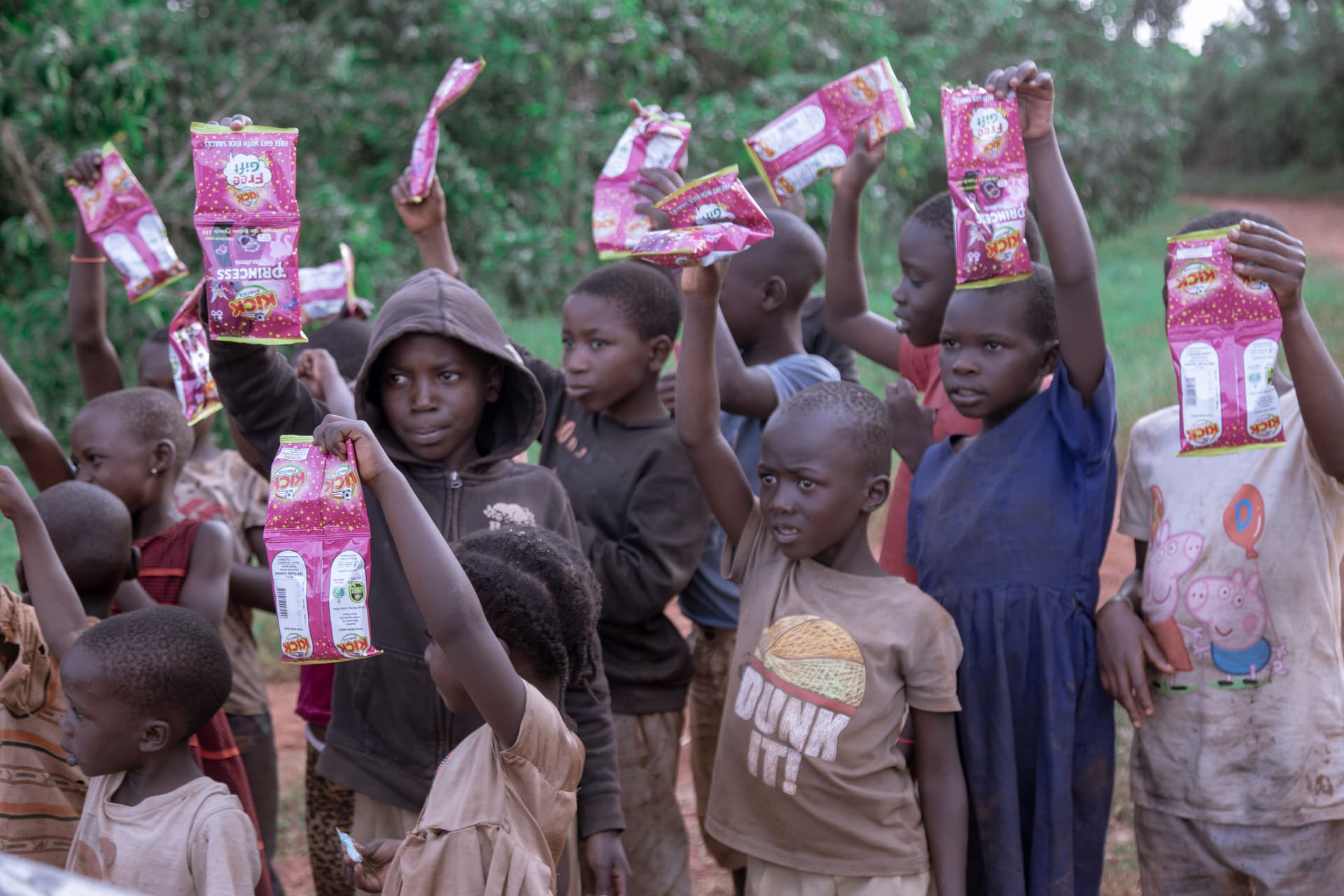 Group of children and adults standing outdoors, some holding boxed food packages overhead.