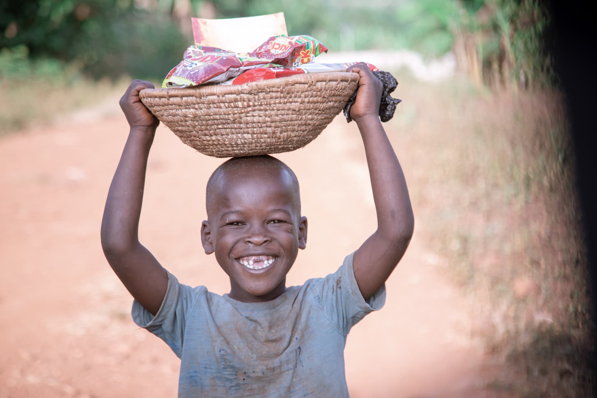 Boy smiling while balancing a basket of food and blankets on his head outdoors.