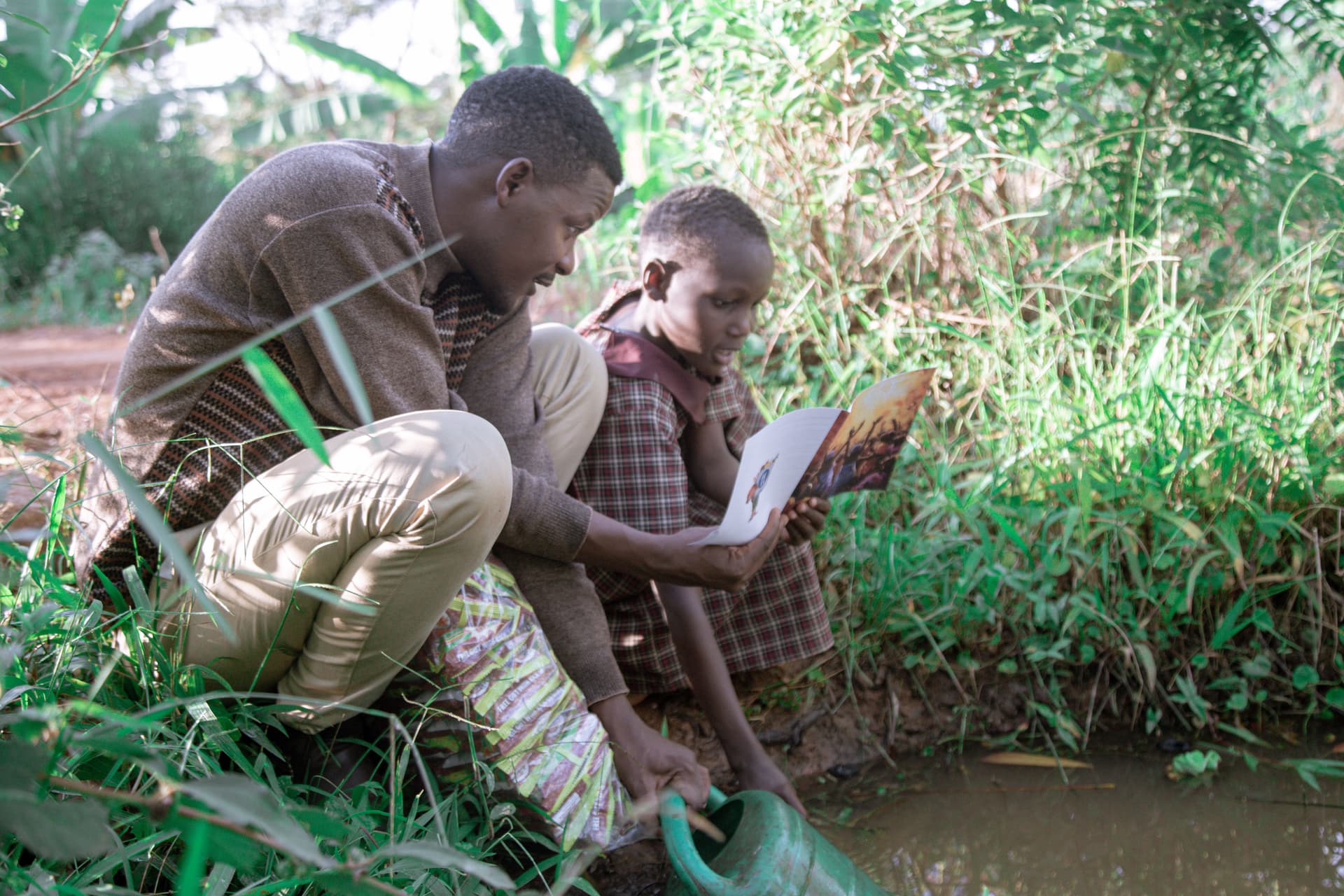 Man kneeling beside a seated elderly man, reading a document together near a stream.