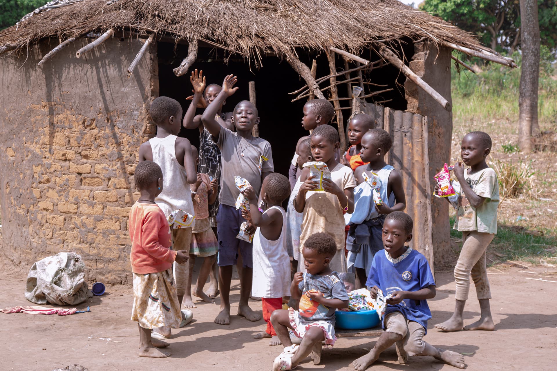 A group of children gathers under a thatched shelter, several waving toward the camera.