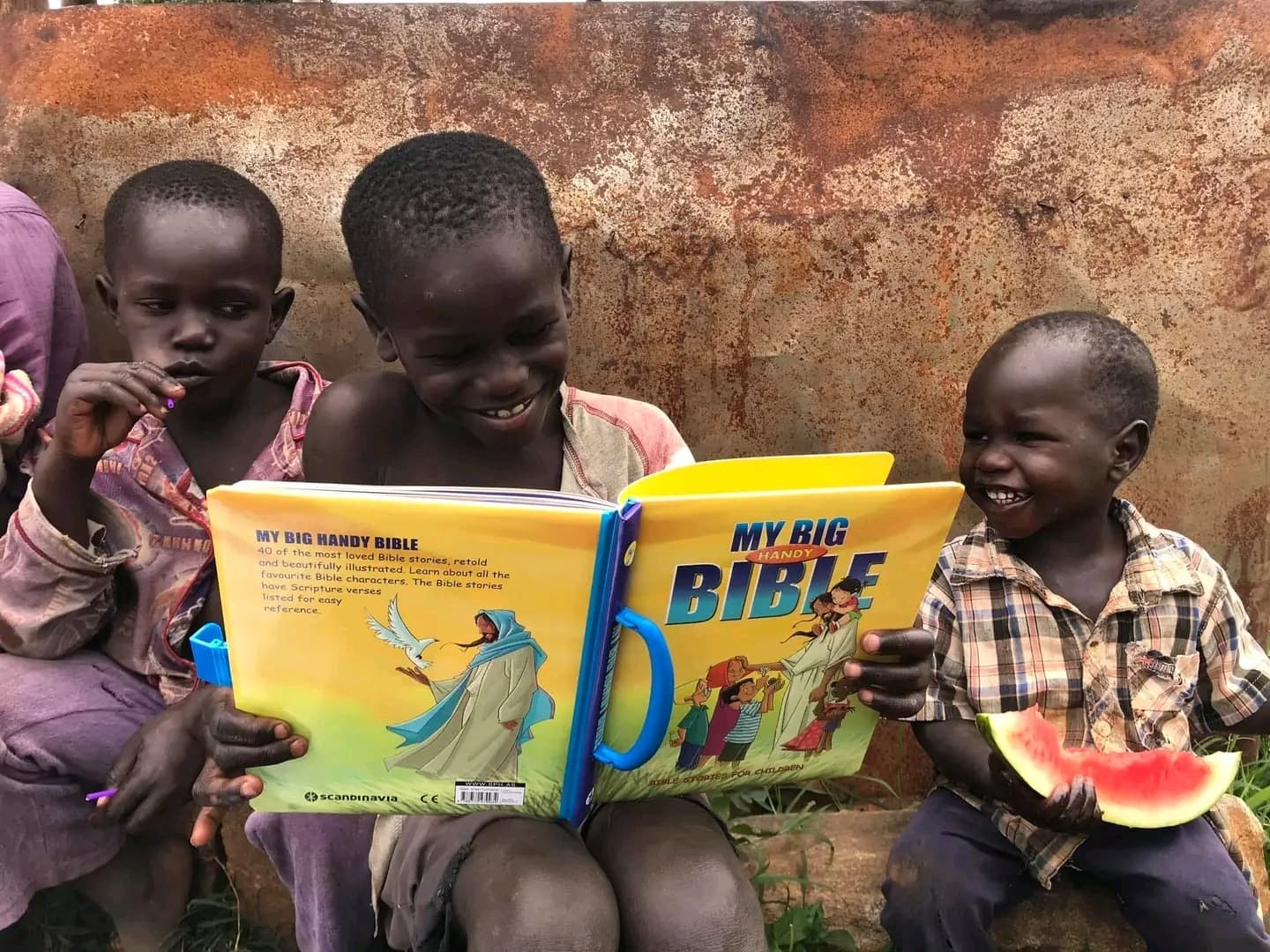 Two children sit outdoors reading a colorful children's Bible together, one eating watermelon.