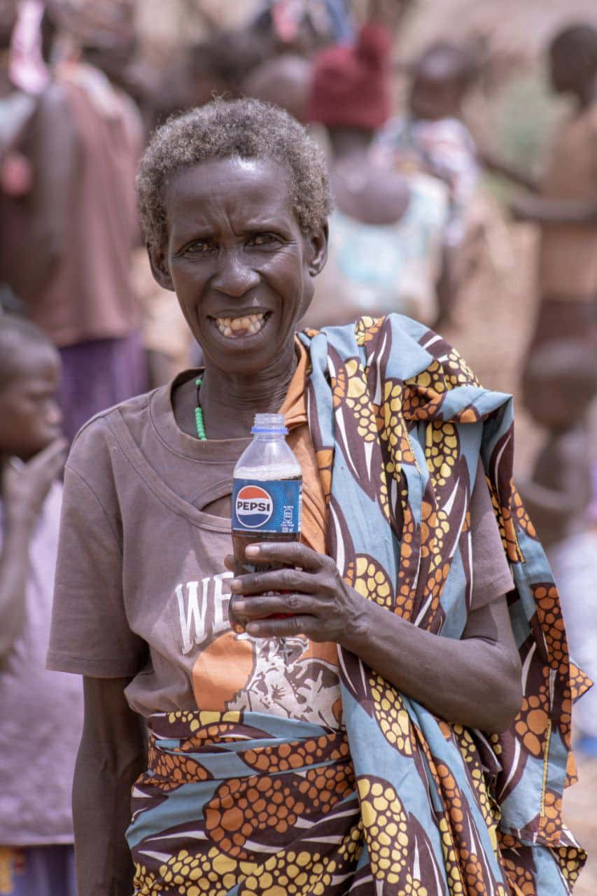 Elder Elizabeth, an elderly woman in a patterned dress, smiles while holding a small bottle, crowd behind her.