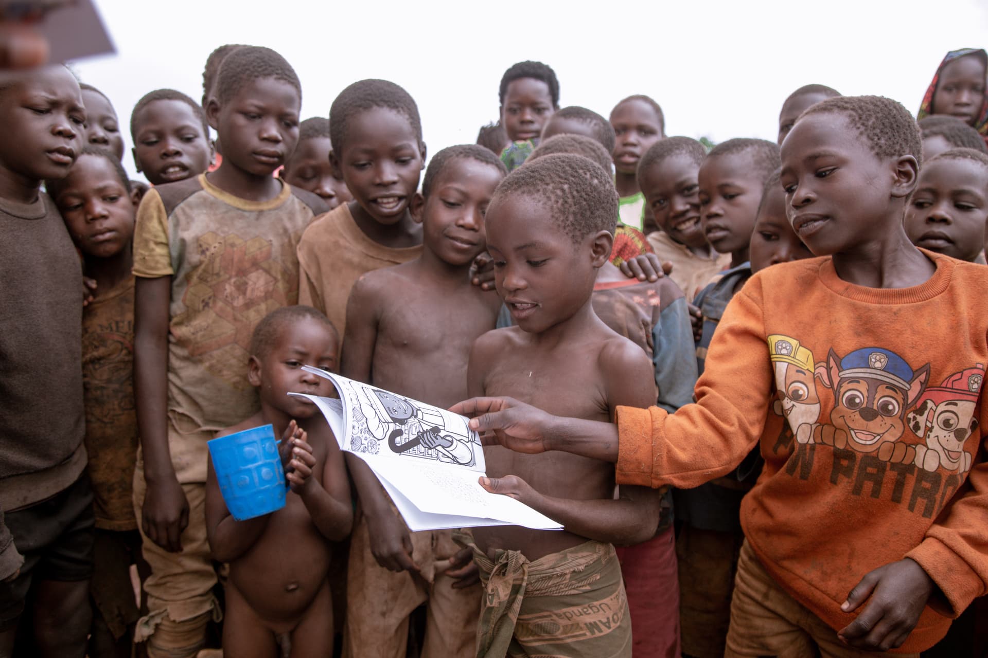 Children in orange clothing looking closely at an open coloring book page, one holding a blue cup.
